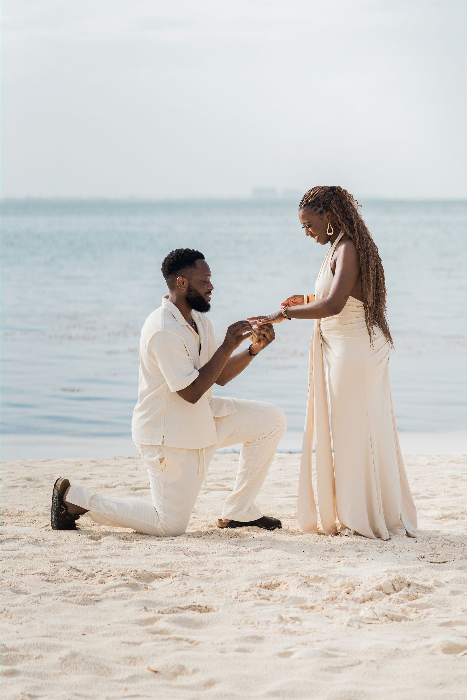 Couple sharing romantic beach proposal moment, man on knee placing ring on womans finger by the sea.