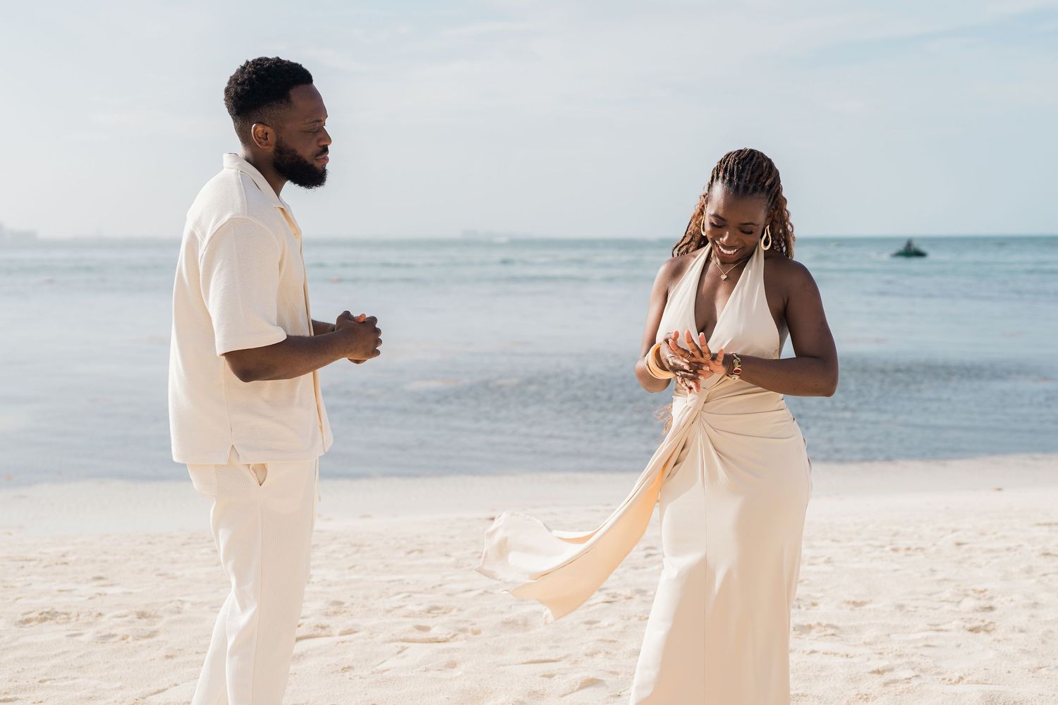 Happy couple dancing on a white sand beach in matching white outfits with calm ocean behind them.