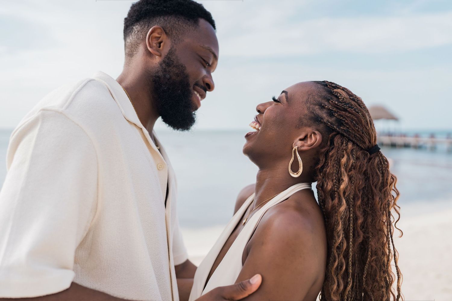 A joyful Black couple sharing a romantic moment on a sunny beach, smiling at each other near a pier.