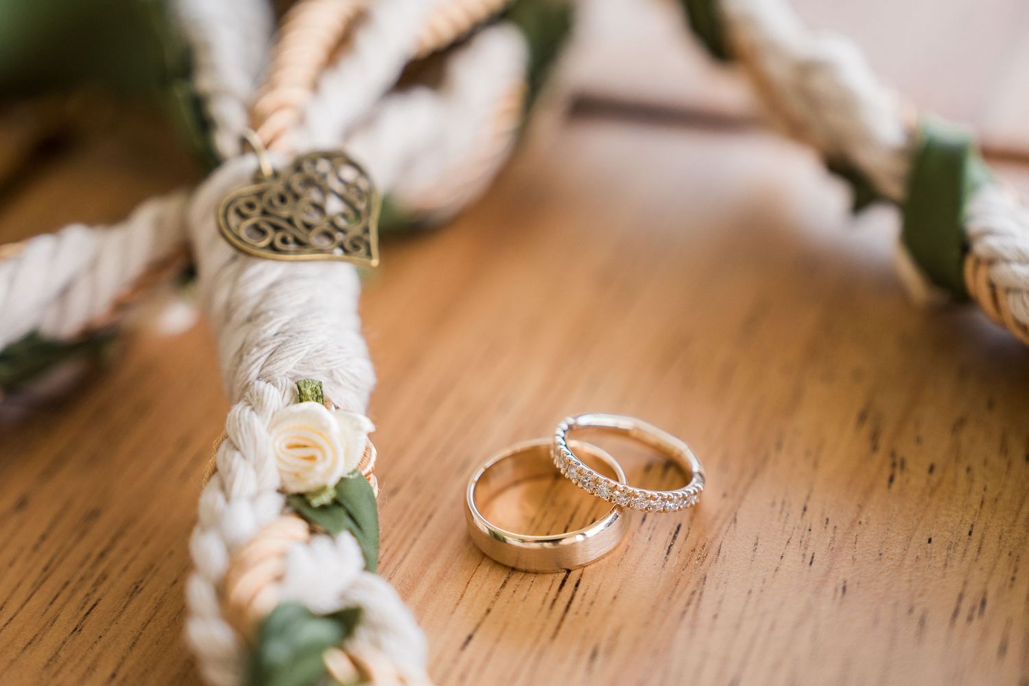 Hotel Xcaret México Wedding Two gold wedding rings rest on a wooden surface beside a floral rope decoration with white flowers and greenery.