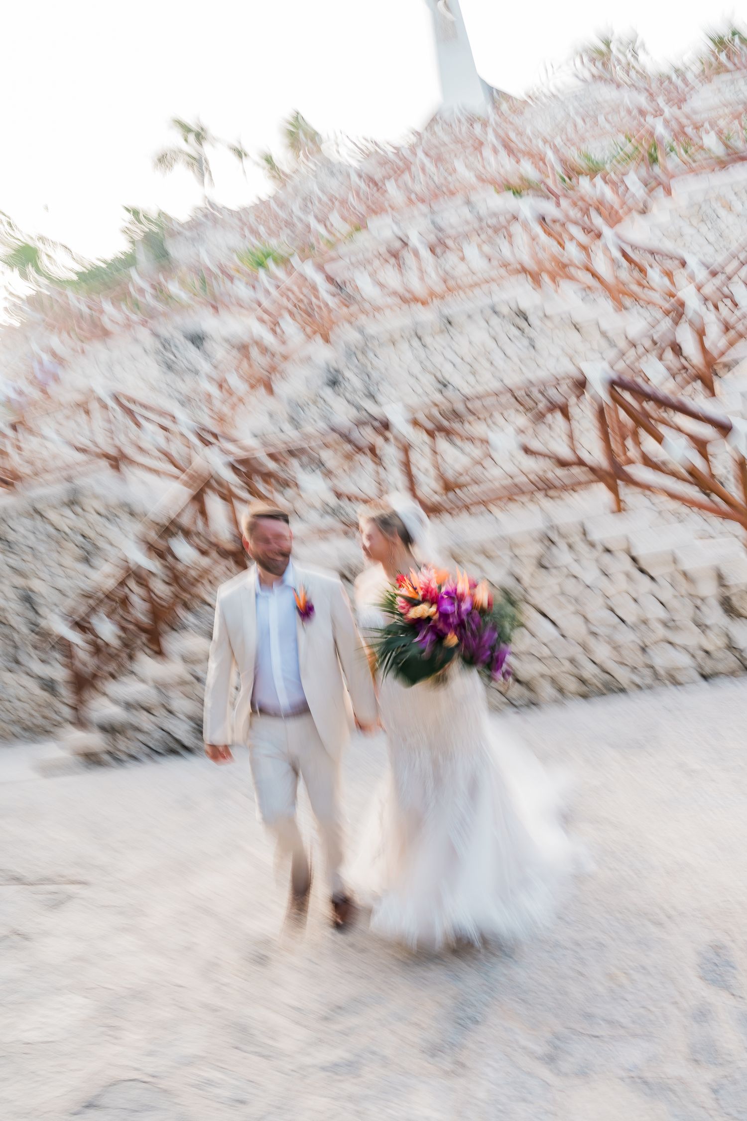 Hotel Xcaret México Wedding - Bride and groom walking hand-in-hand on sandy beach, bride holding colorful bouquet, motion blur effect.