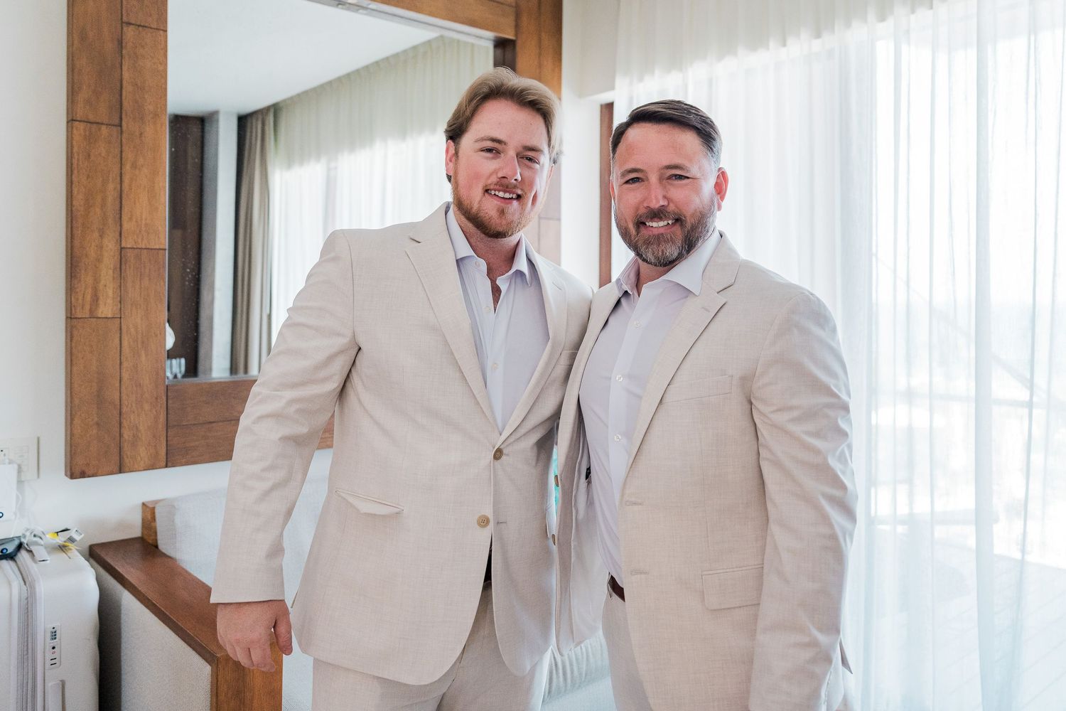 Two men in matching beige suits smiling together in a bright, elegant room with white curtains.