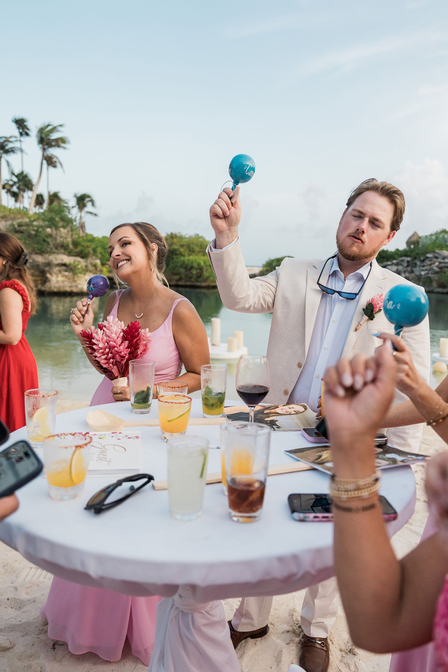 Hotel Xcaret México Guests shaking blue maracas at a tropical beach wedding reception table with colorful drinks.
