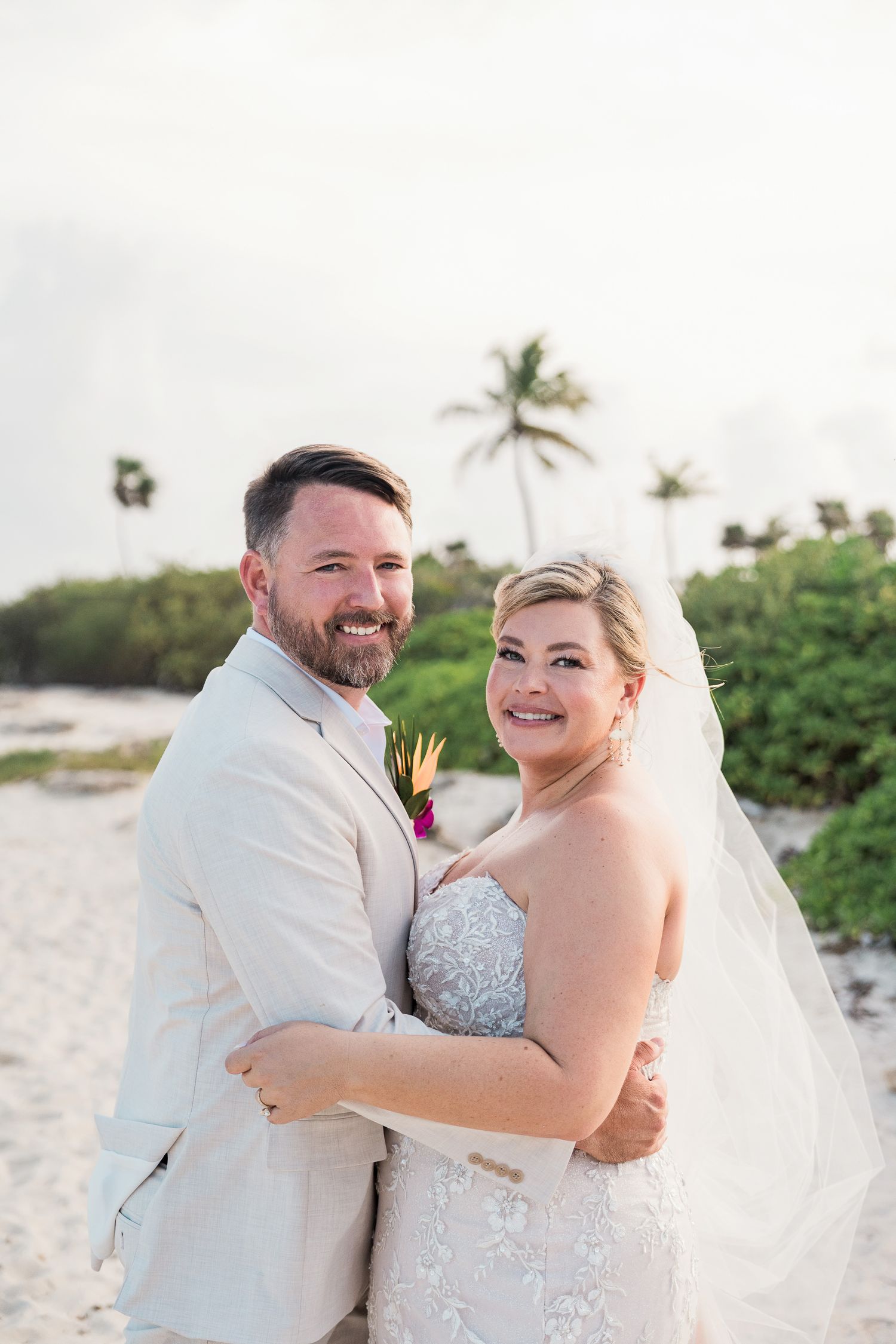 Bride and groom embrace on a tropical beach, smiling, groom in tan suit, bride in lace gown with veil, palm trees behind.