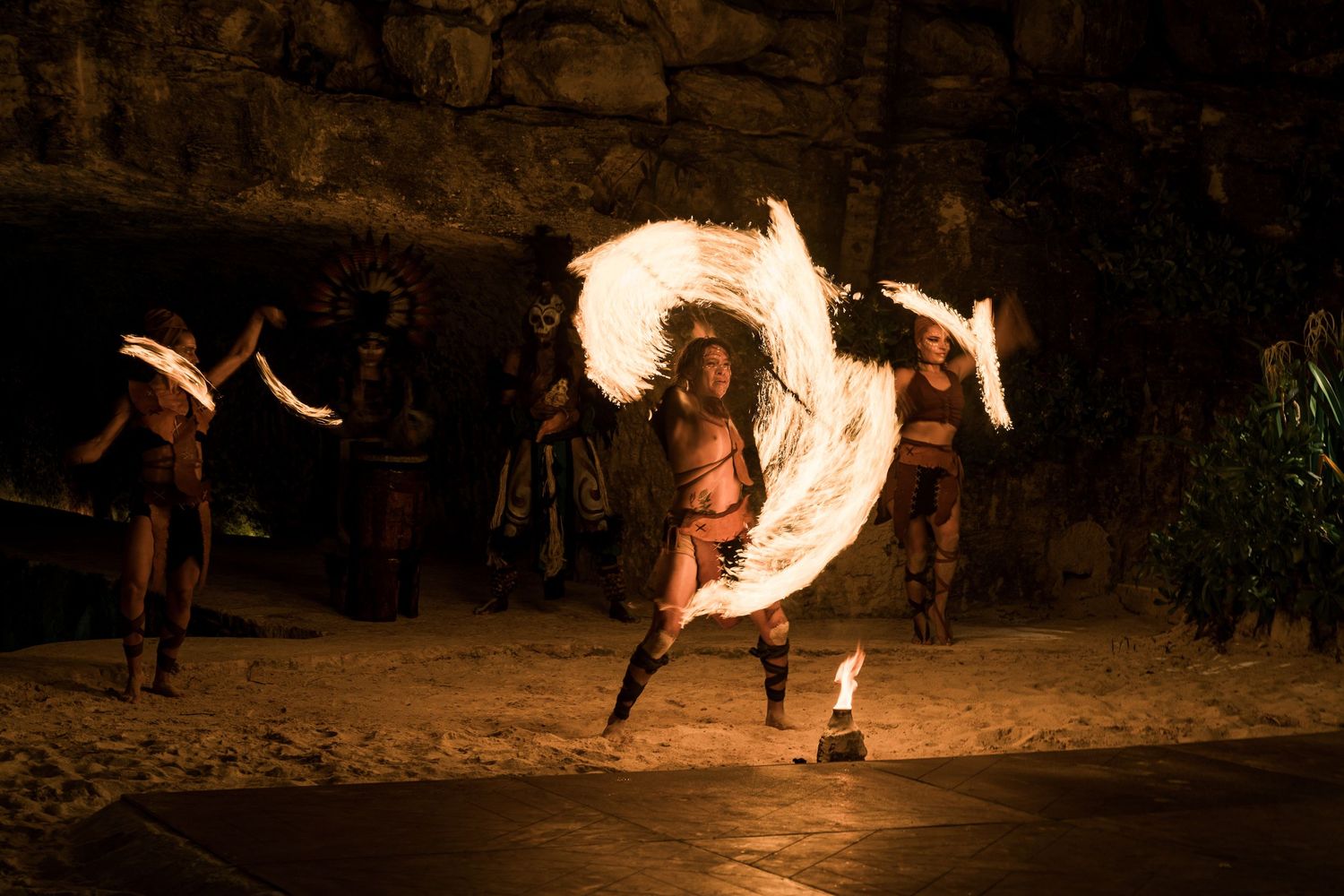 Hotel Xcaret México Fire dancers perform with spinning flames in a dark cave setting on sandy ground at night.
