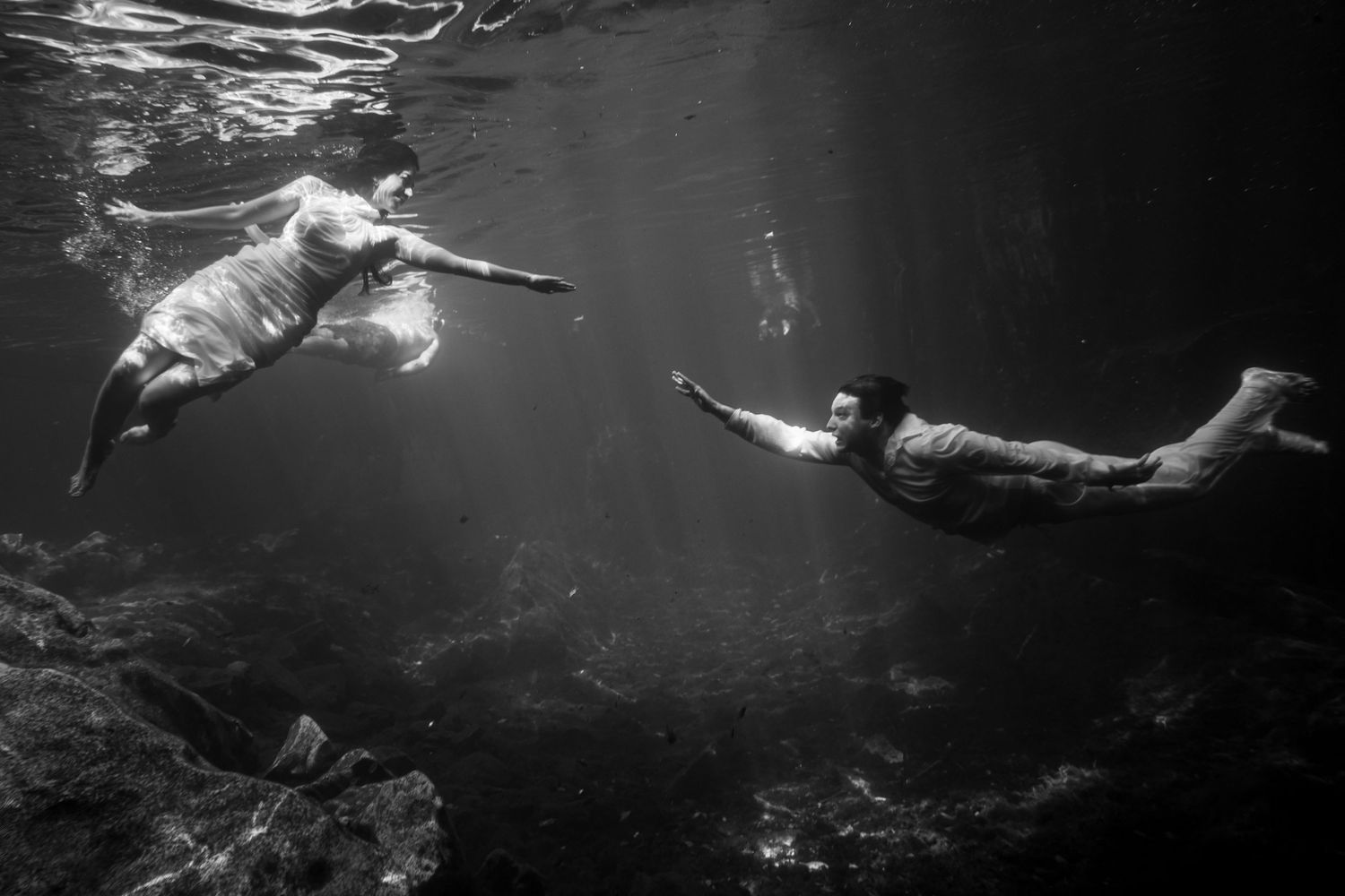 Black and white underwater photo of two people swimming toward each other in a sunlit cenote.