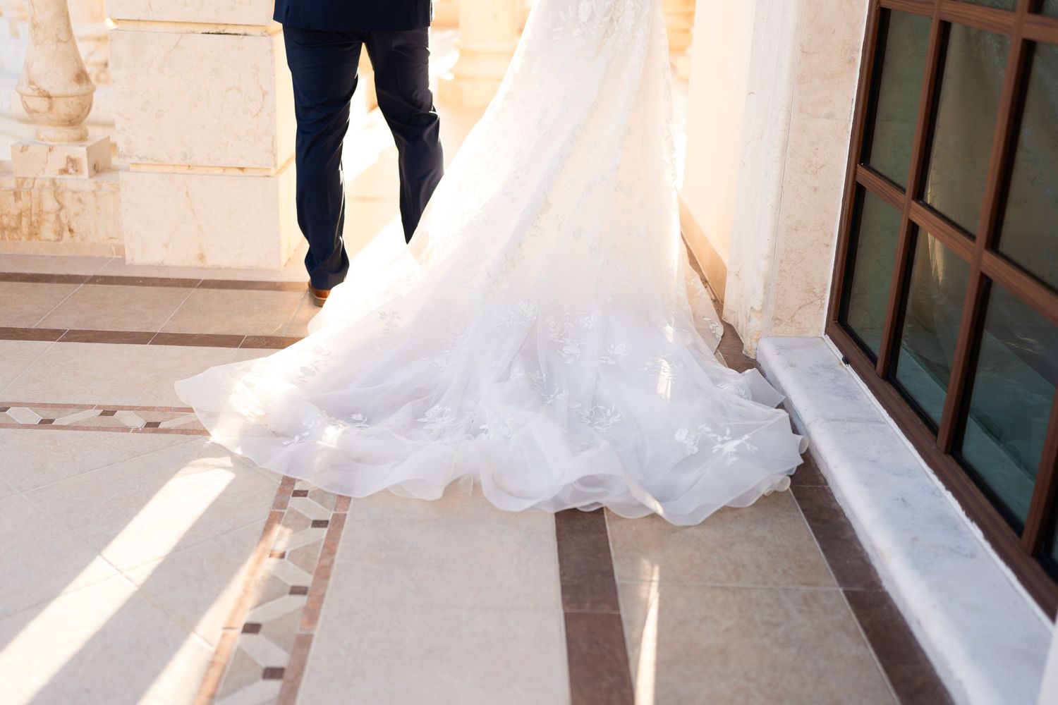 Detail shot of flowing white wedding gown train on marble floor near ornate building.