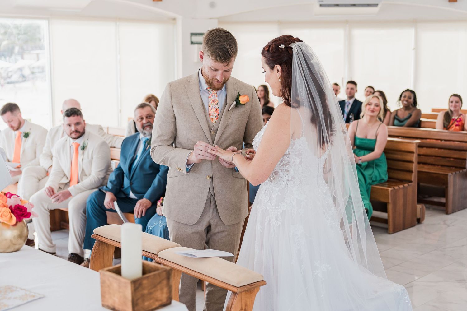 Groom places wedding ring on bride's finger during an intimate chapel ceremony with guests watching.