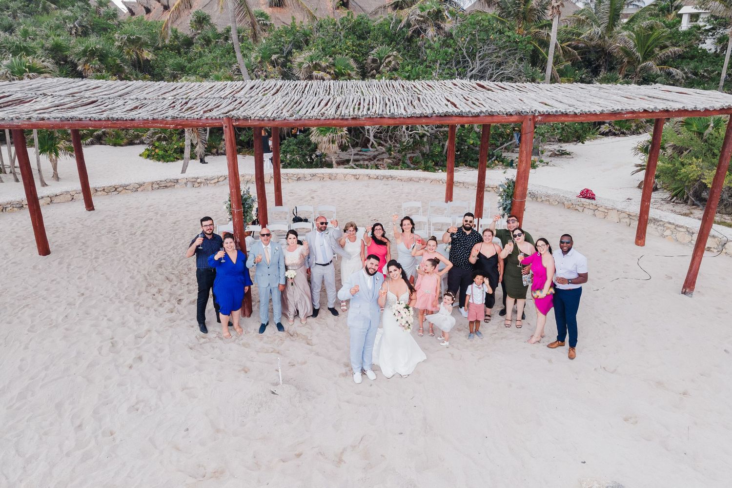 Aerial view of wedding party posing together under a thatched pergola on a white sandy beach.