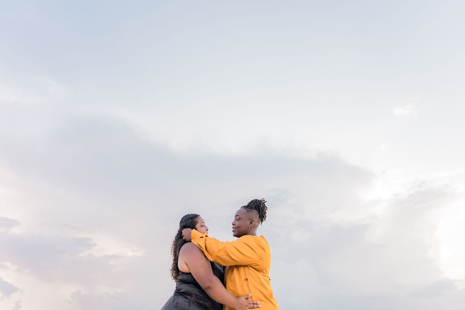 Two women embracing against a soft cloudy sky at sunset.
