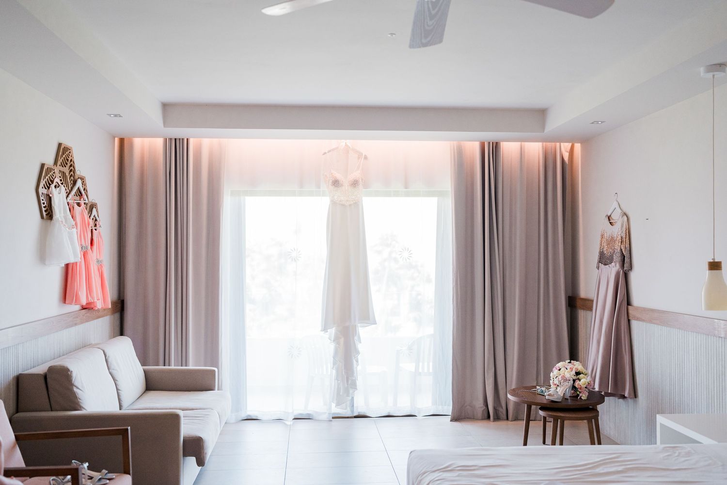 Wedding dress hanging in bright hotel room with pink curtains, sofa, and bridal party attire.
