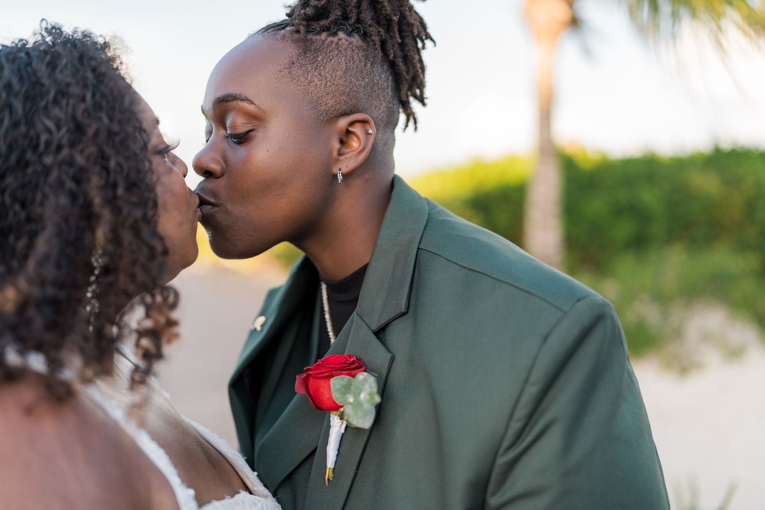Newlyweds share a romantic kiss on the beach with palm trees in background.