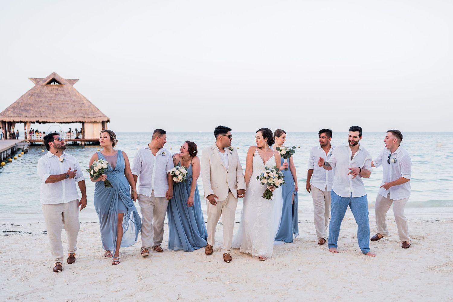 Wedding party walks together on a sandy beach at sunset with a thatched pier in the background.