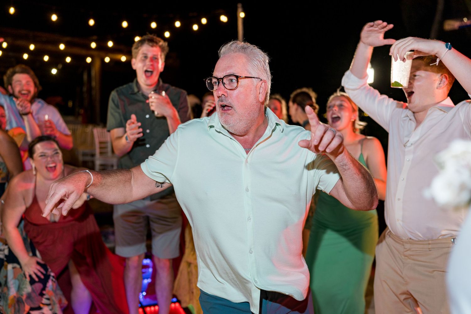 Man in mint shirt dancing joyfully with wedding guests under string lights.