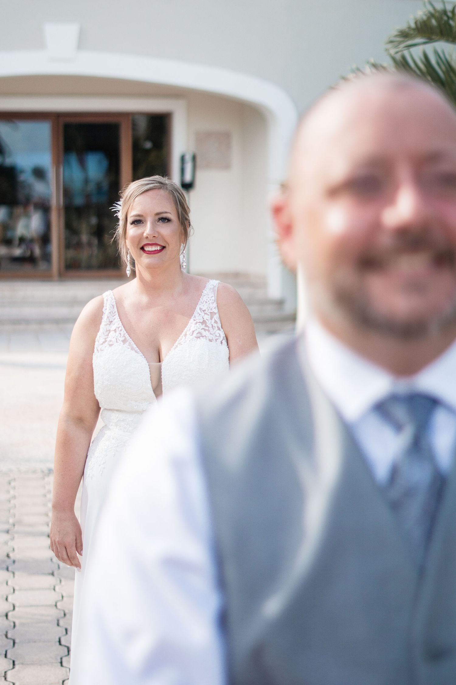 Smiling bride in lace V-neck wedding dress approaches groom in gray vest during first look.