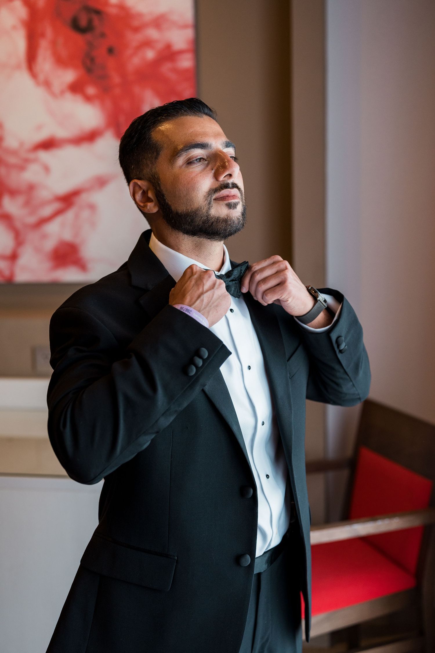Handsome groom in black tuxedo adjusts bow tie while getting ready for wedding.