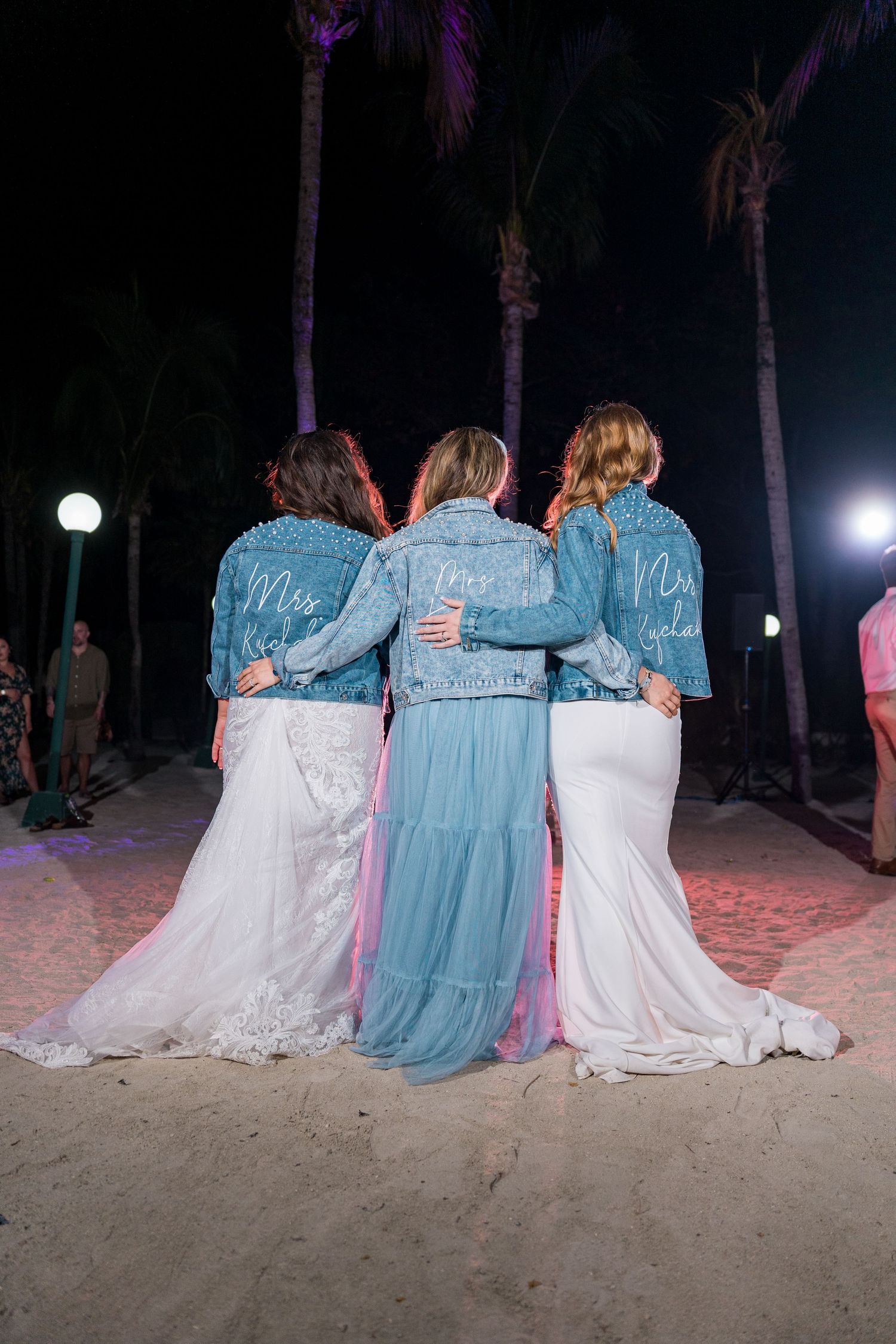 Three women in pearl-studded denim bridal jackets and white gowns pose on a beach at night under palm trees.