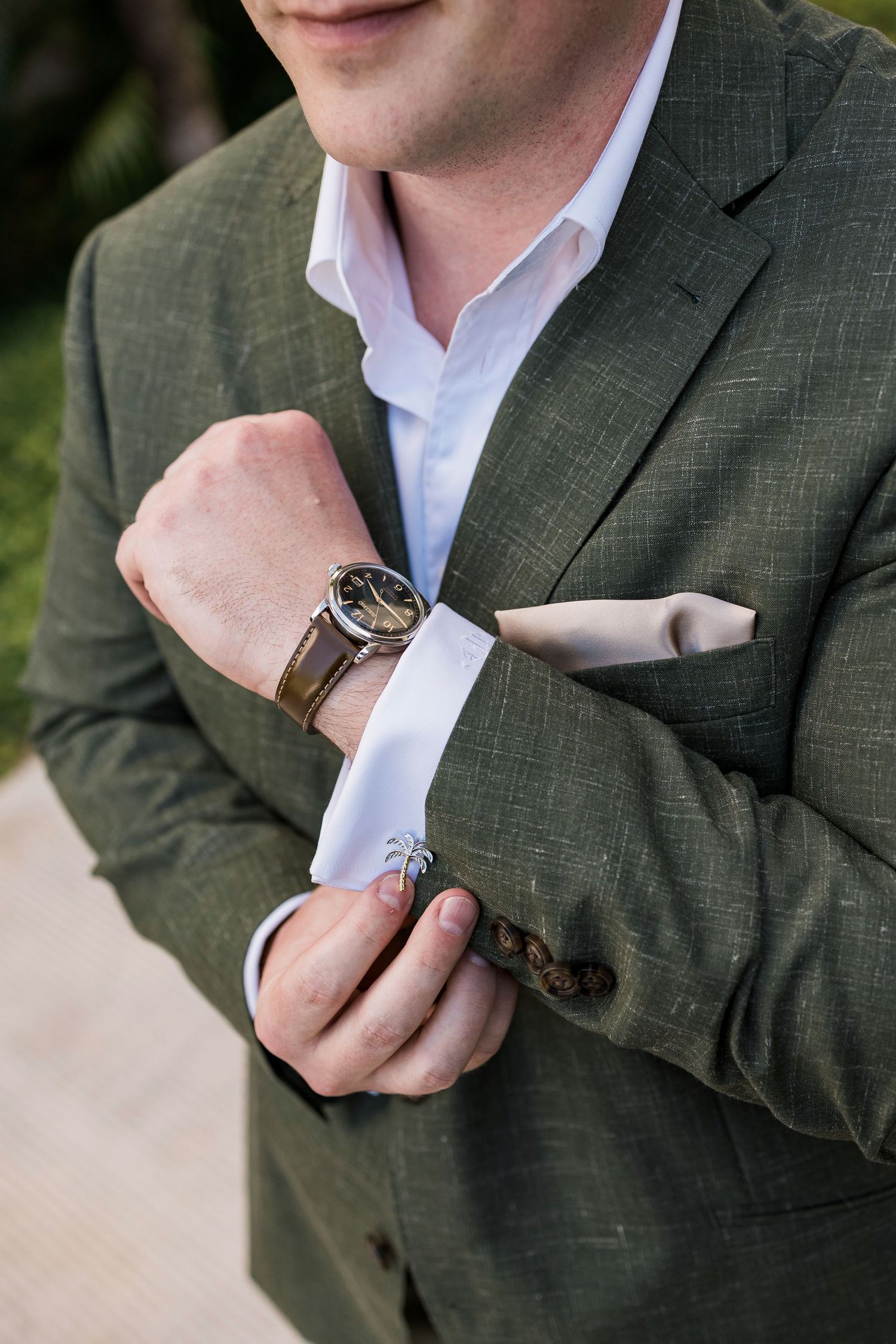 Man in olive green suit adjusting cufflinks on white dress shirt, wearing a brown leather watch.