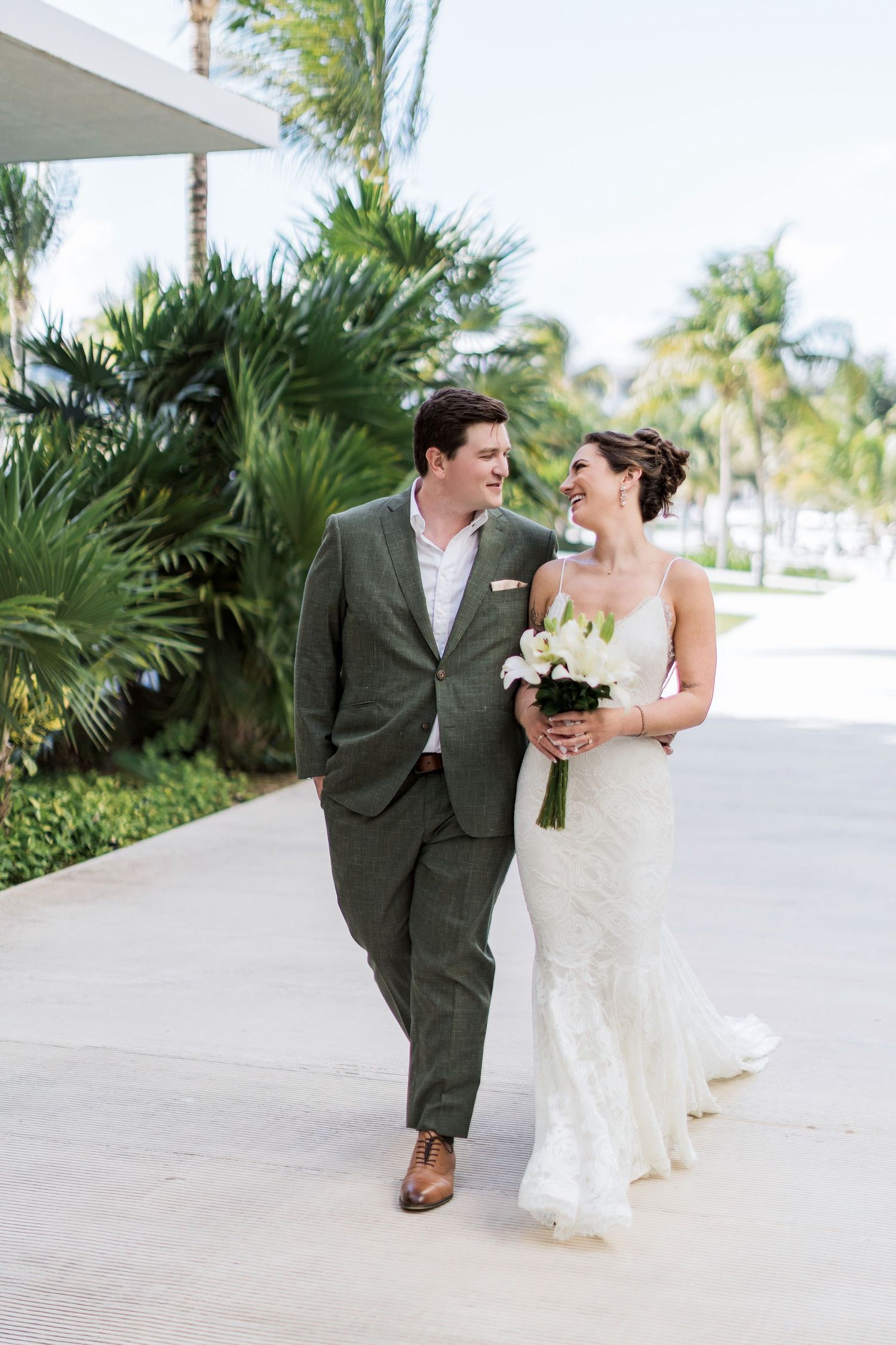Bride and groom walking together outdoors at tropical wedding venue, bride in lace gown holding white bouquet.