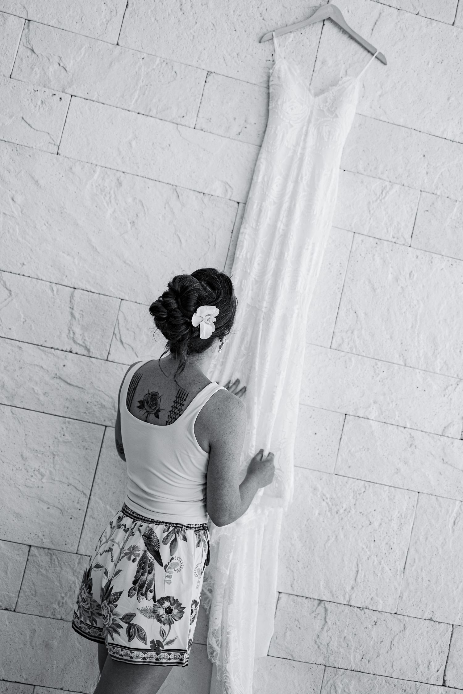 Bride in casual attire admiring her wedding dress hanging on wall in black and white photo.