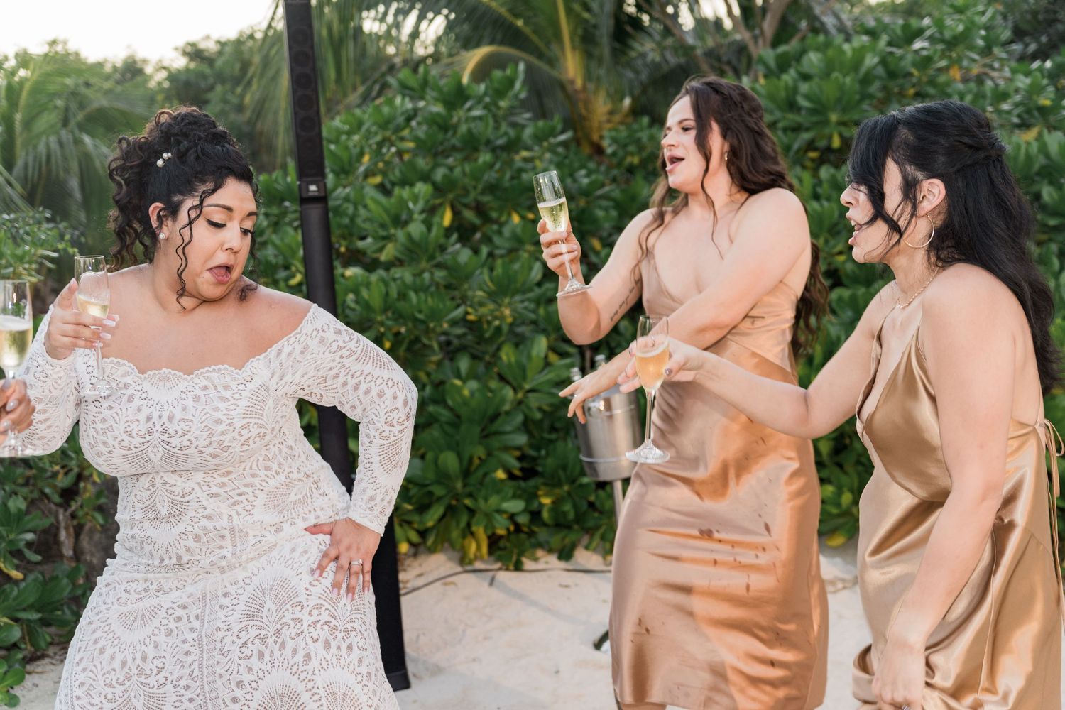 Xcaret Arte Wedding - Bride in lace gown celebrates with bridesmaids in gold dresses, holding champagne glasses outdoors