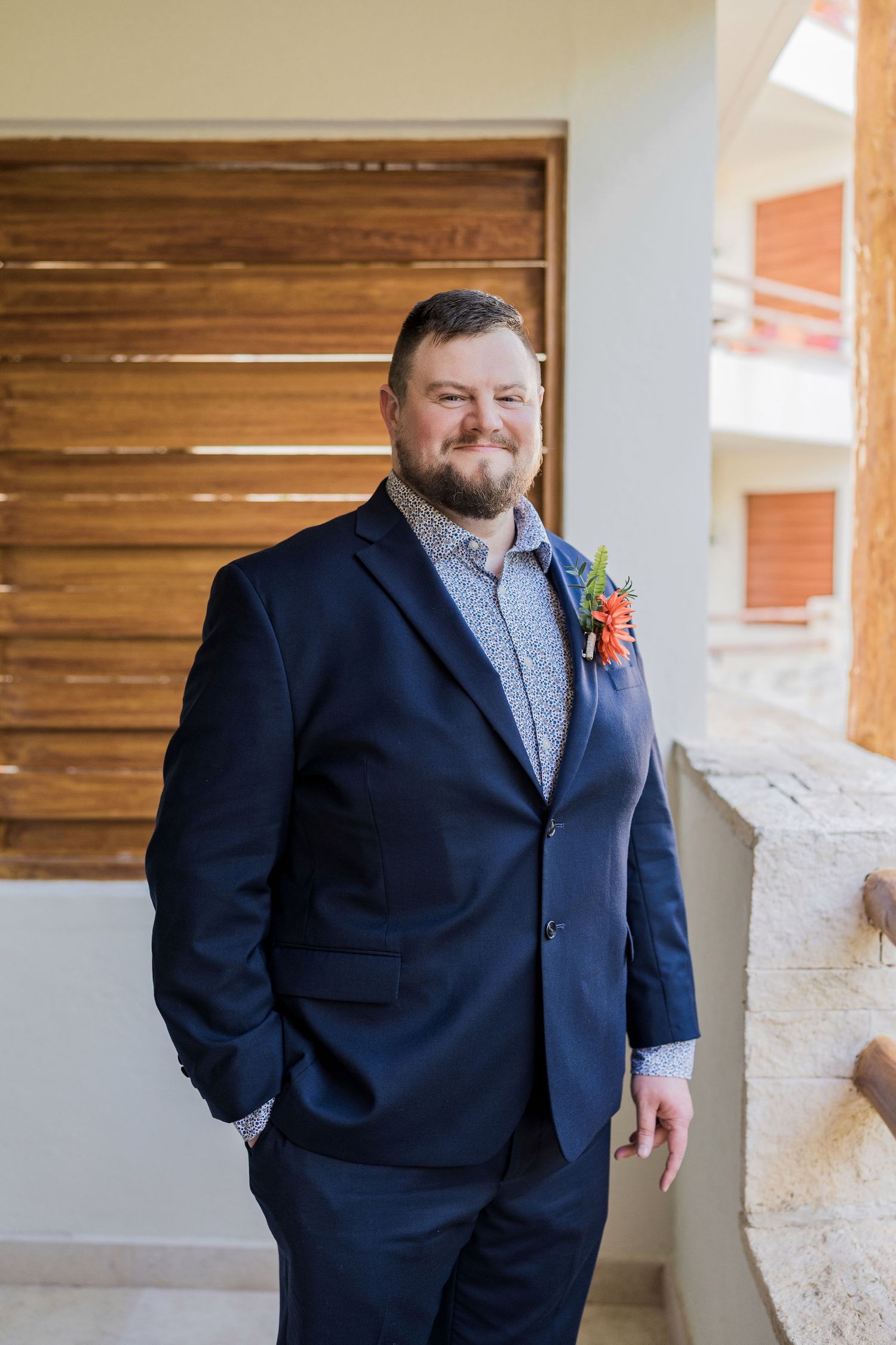 Secrets Akumal Wedding - A smiling groom in a navy blue suit with a colorful boutonniere stands on a balcony at a destination wedding.
