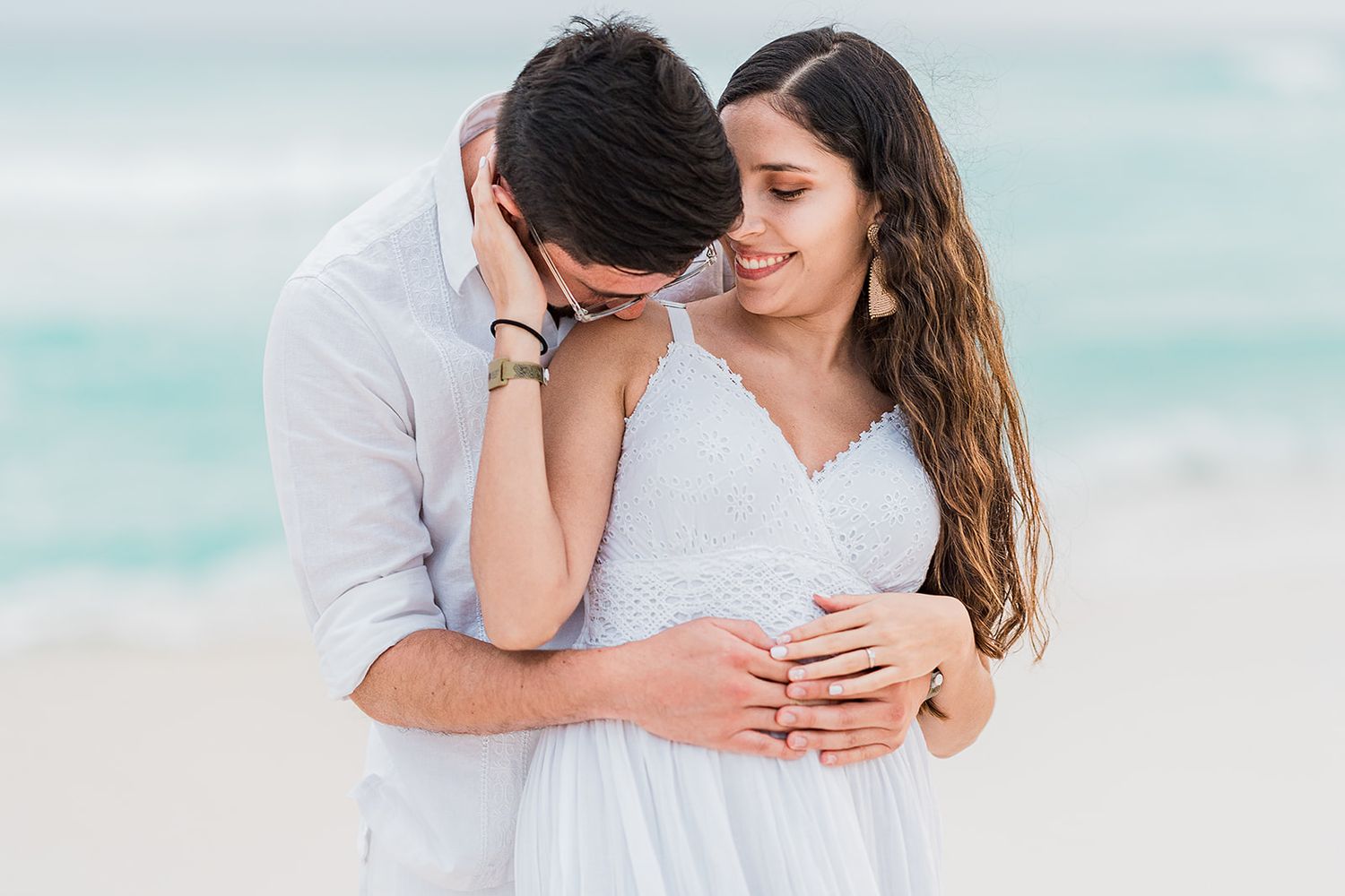 Romantic couple embracing on a tropical beach, woman in white dress, man in white shirt, ocean waves in background. Cancun Couple Photoshoot