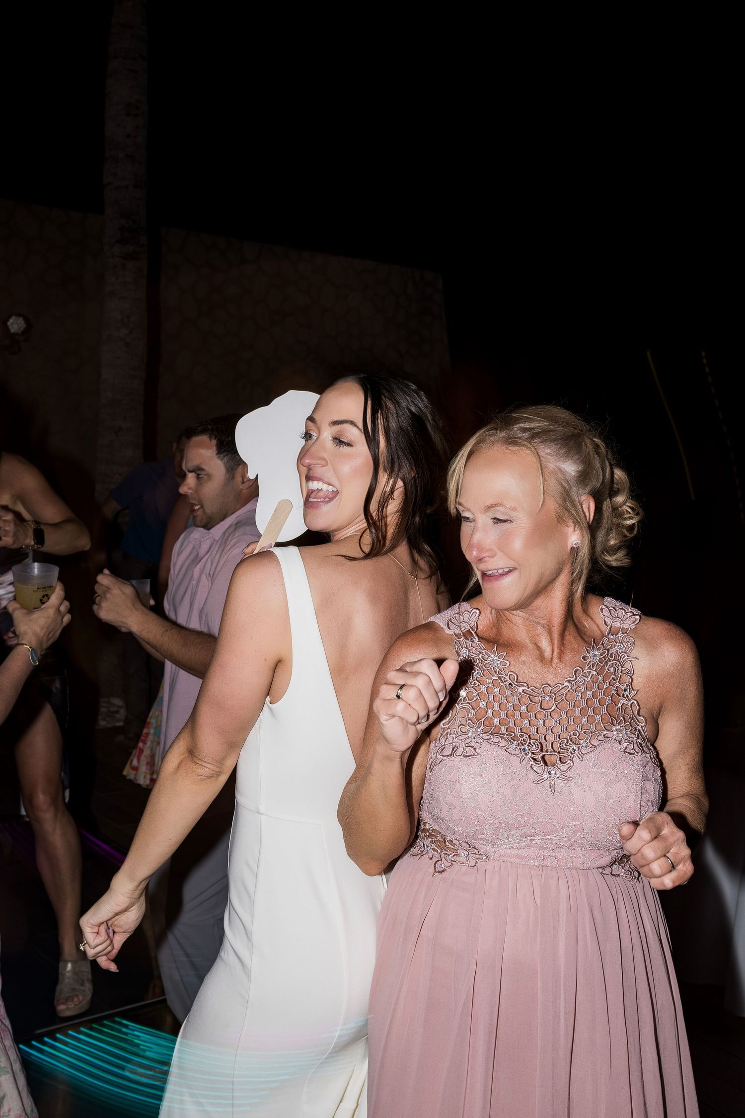 Bride in white dress and older woman in pink gown dancing joyfully together at an outdoor wedding reception.