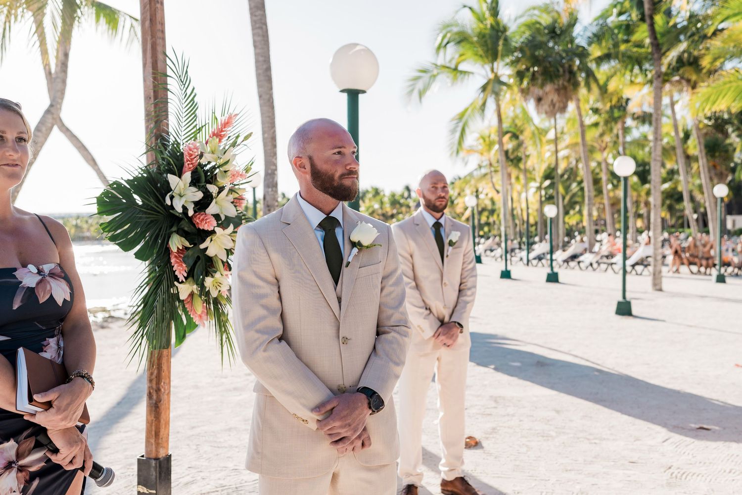 Groom in beige suit stands at beach wedding altar with palm trees and floral arrangement under sunny sky.