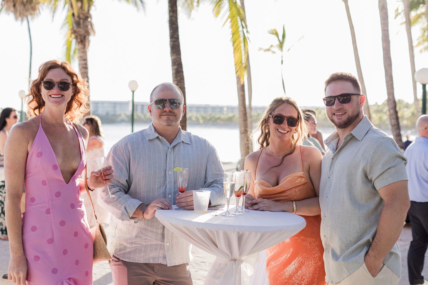 Four friends smiling at an outdoor beachside cocktail party with palm trees and ocean views in a tropical setting.