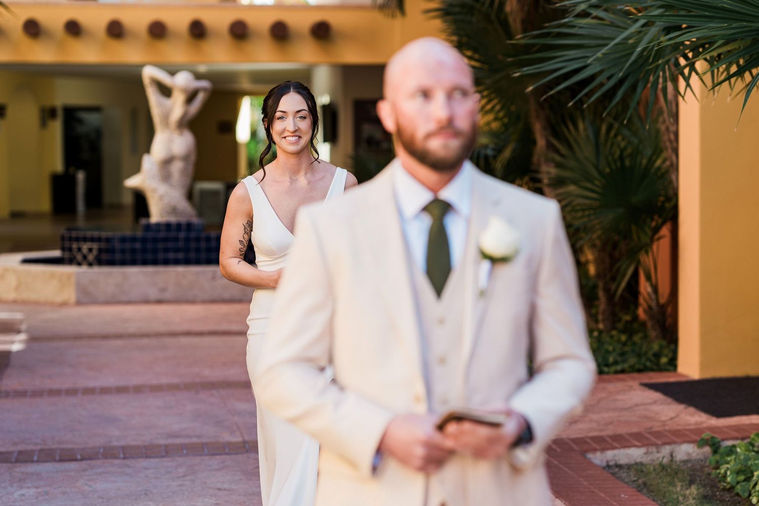 Bride in white gown smiles at groom in cream suit during outdoor wedding first look at a resort.