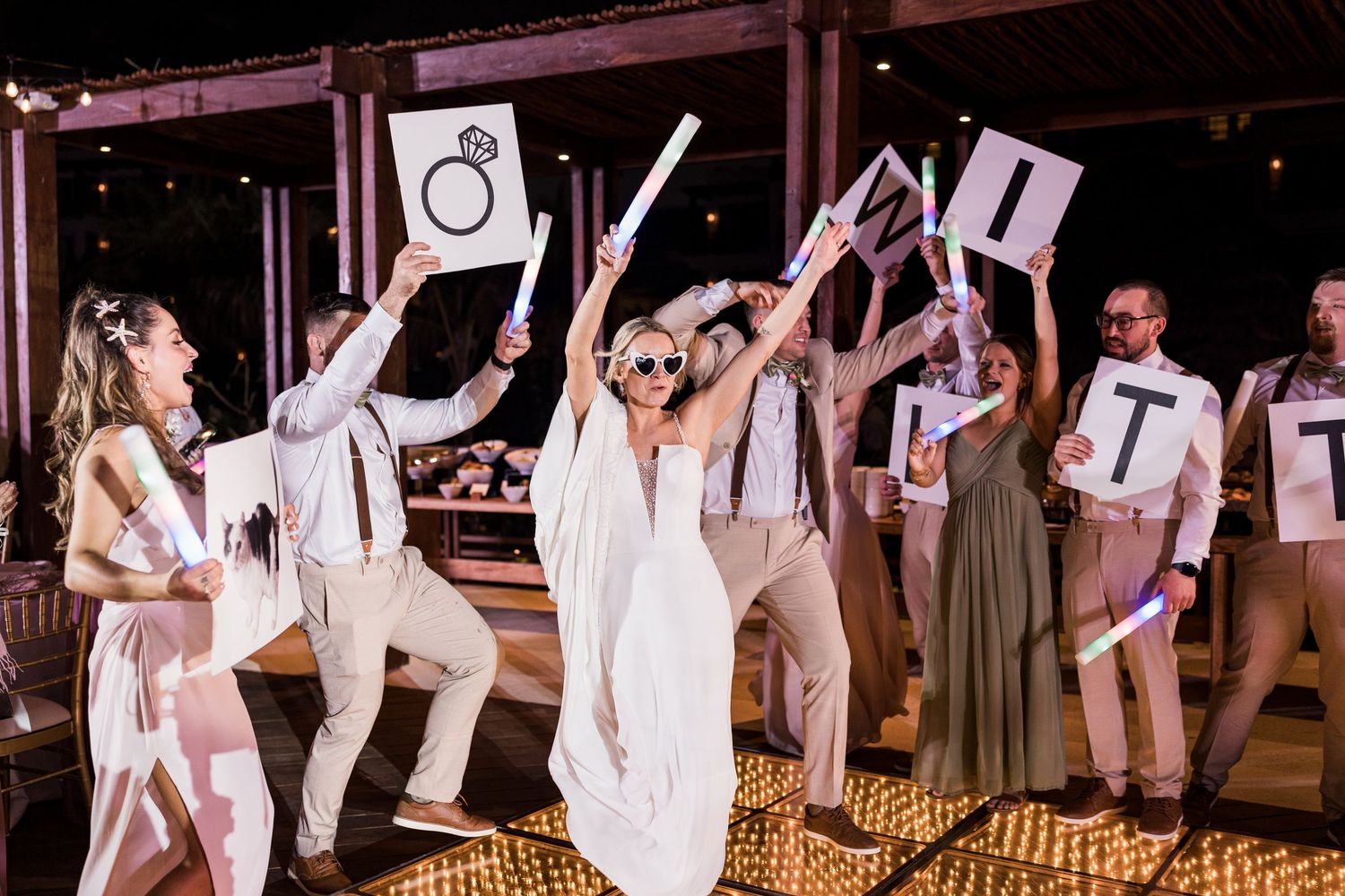 Bride dances joyfully with wedding party holding letter signs on glowing dance floor at lively reception.