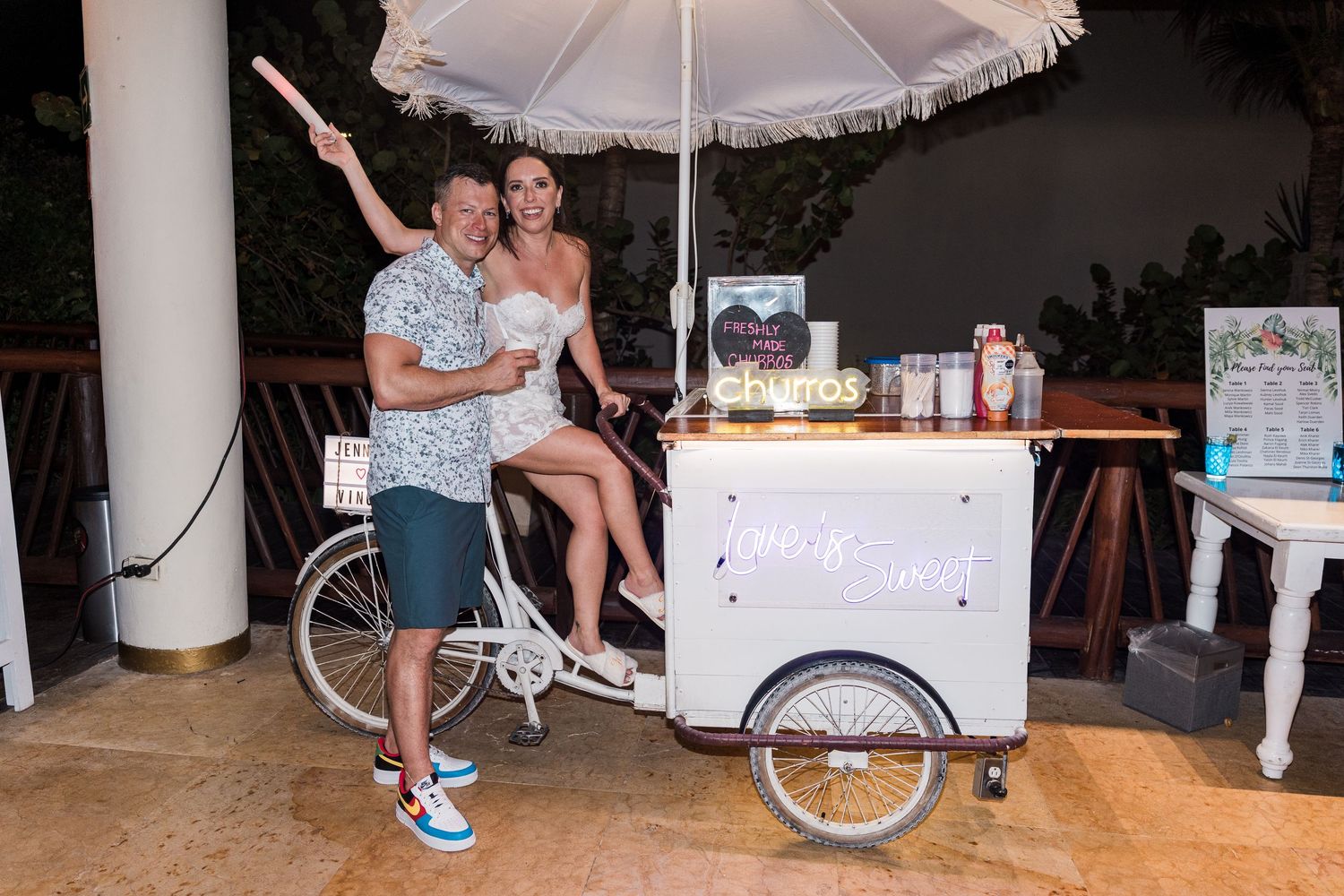 Couple posing with a white ice cream tricycle cart at an outdoor evening event.