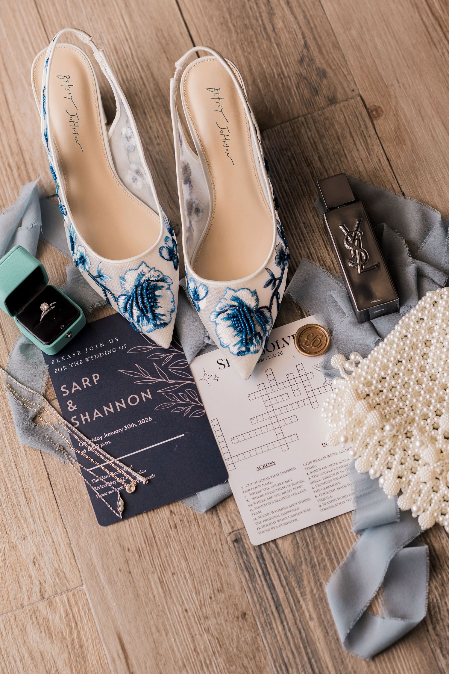 Elegant wedding flat lay with floral embroidered shoes, pearl clutch, YSL perfume, ring box, and navy invitation on wood floor.