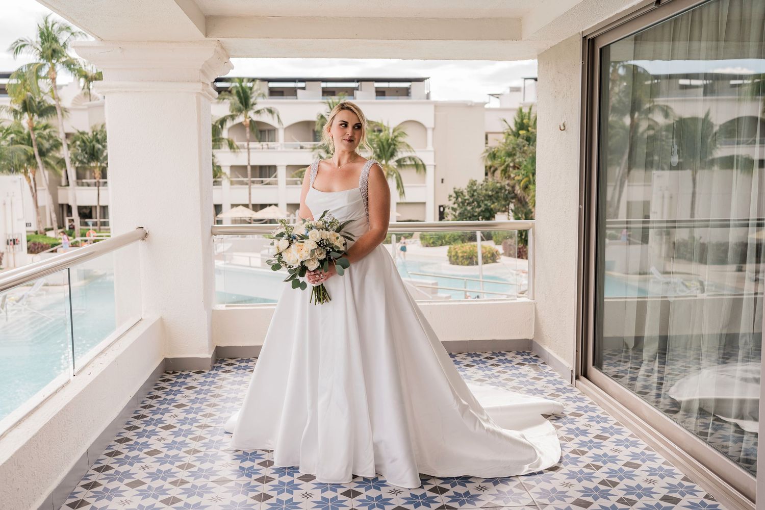 Bride in elegant white ball gown holding bouquet on resort balcony with pool and palm trees in background.