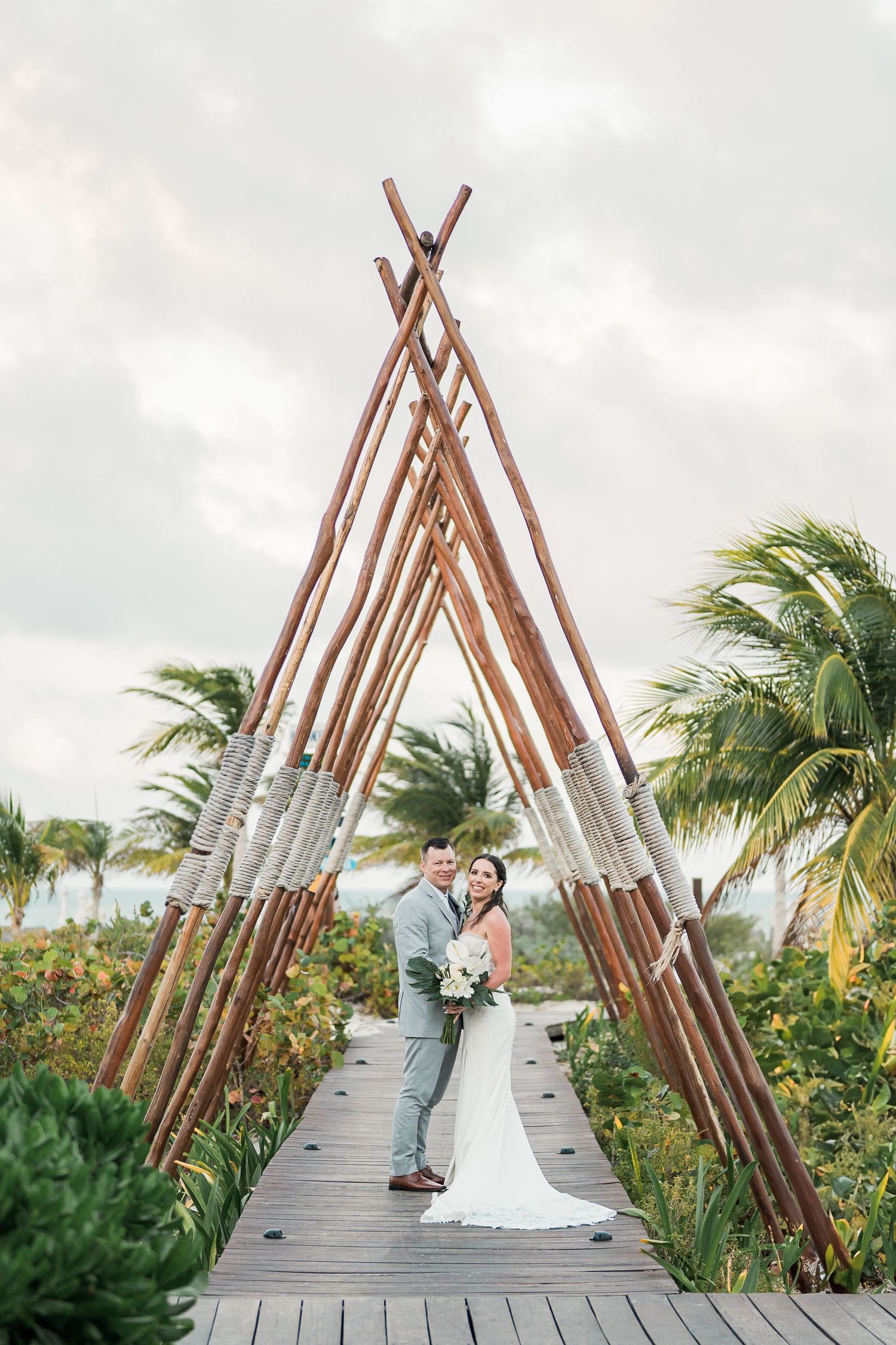 Bride and groom pose under rustic wooden teepee arch on tropical boardwalk in grand palladium costa mujeres