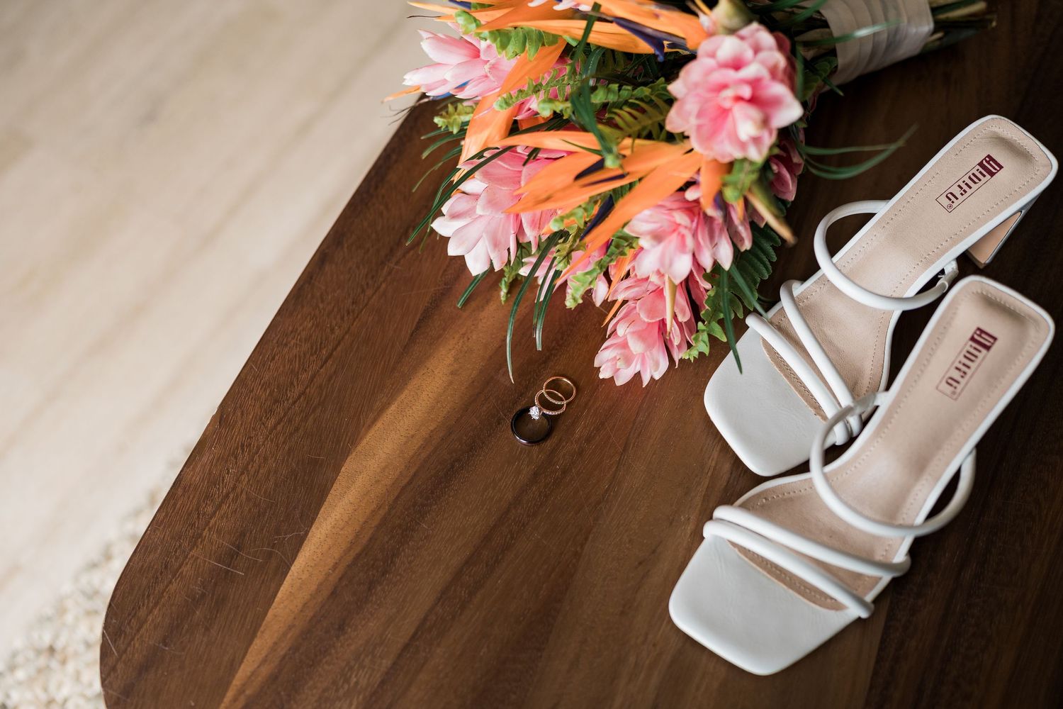 White strappy heeled sandals beside pink and orange flowers and gold rings on a wooden table.