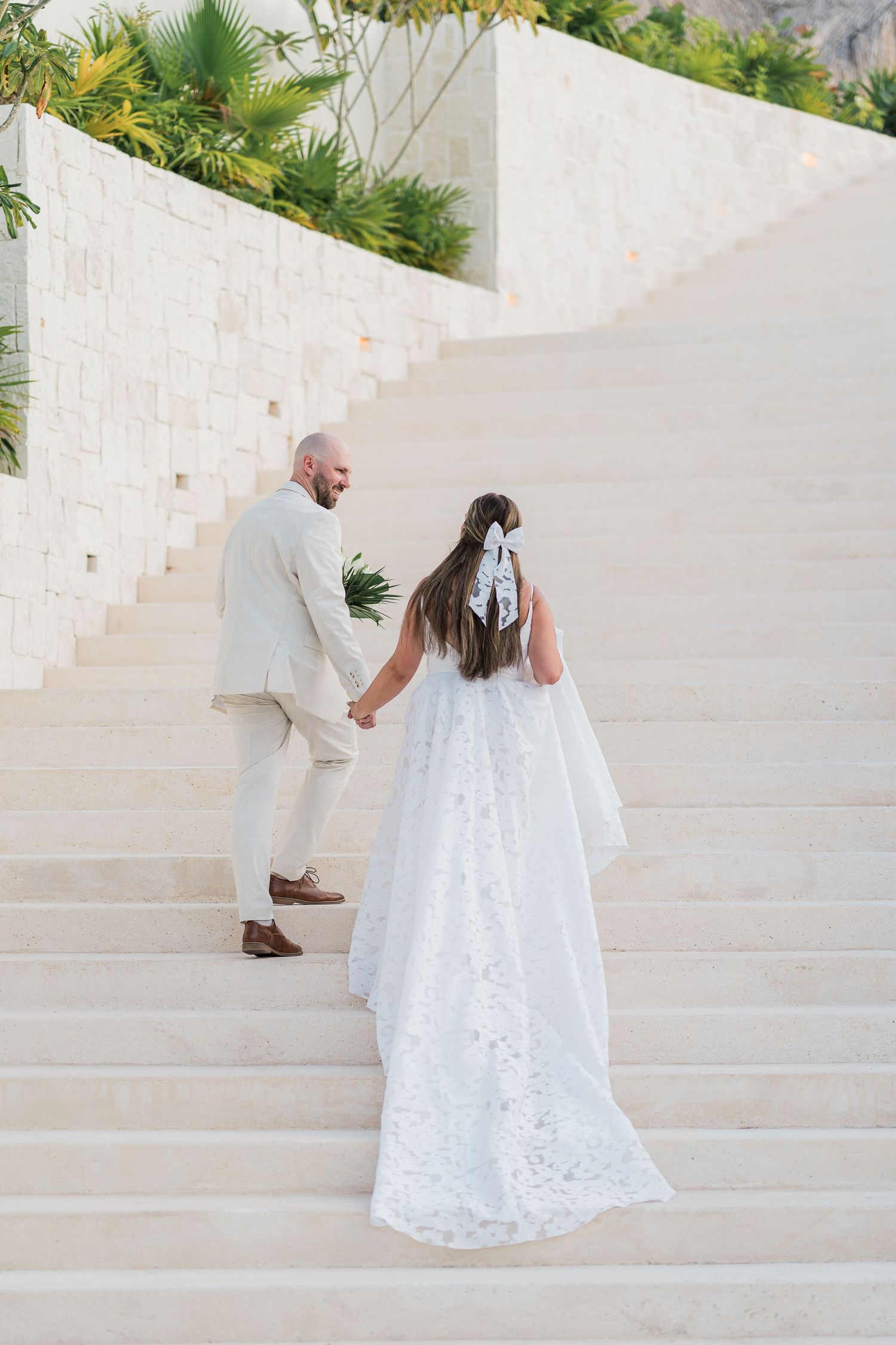 Bride and groom walking up white stone stairs hand in hand, bride wearing lace gown with veil and bow.