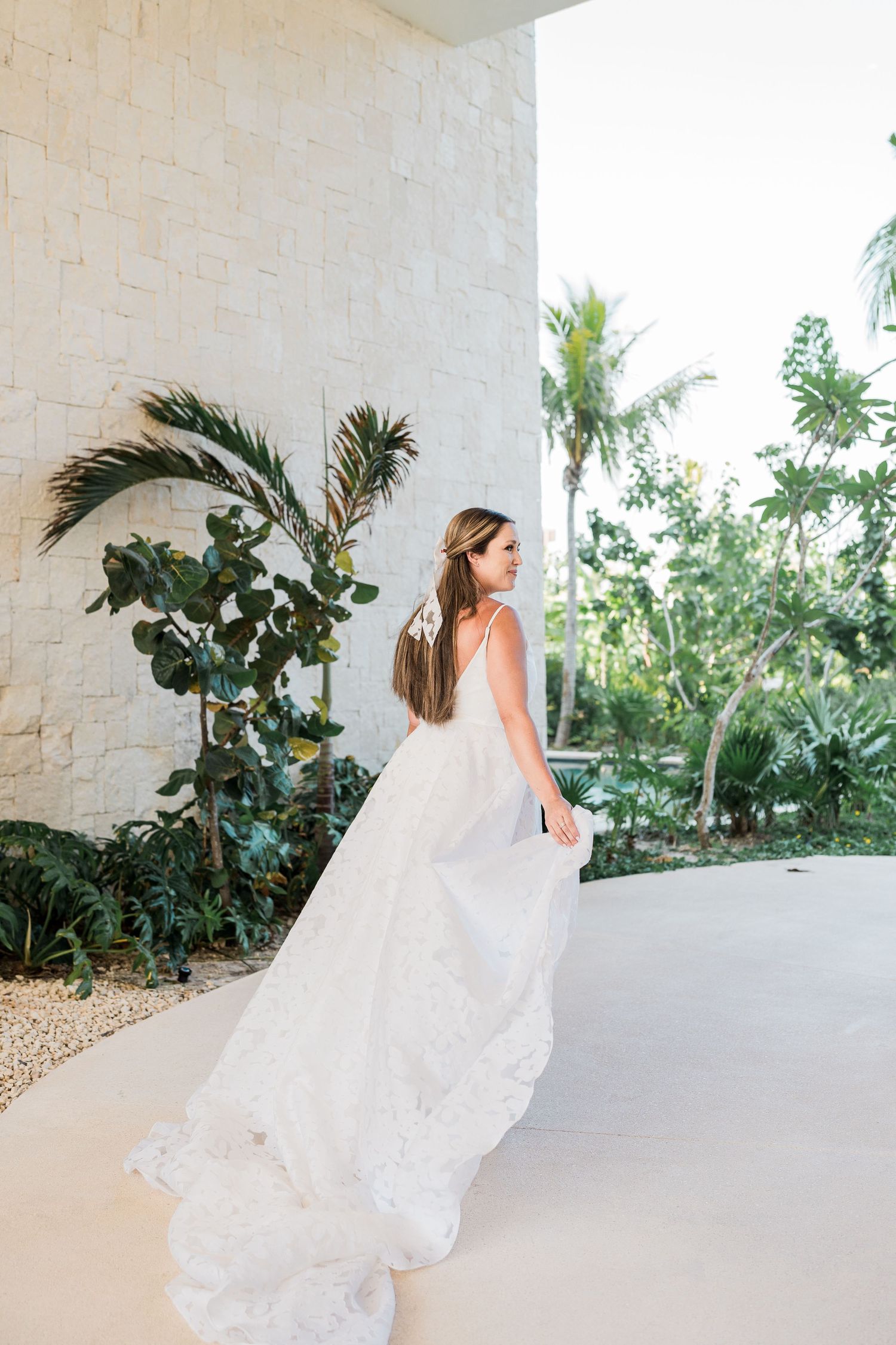 Bride in flowing white lace gown twirling near tropical greenery and palm trees at an outdoor venue.