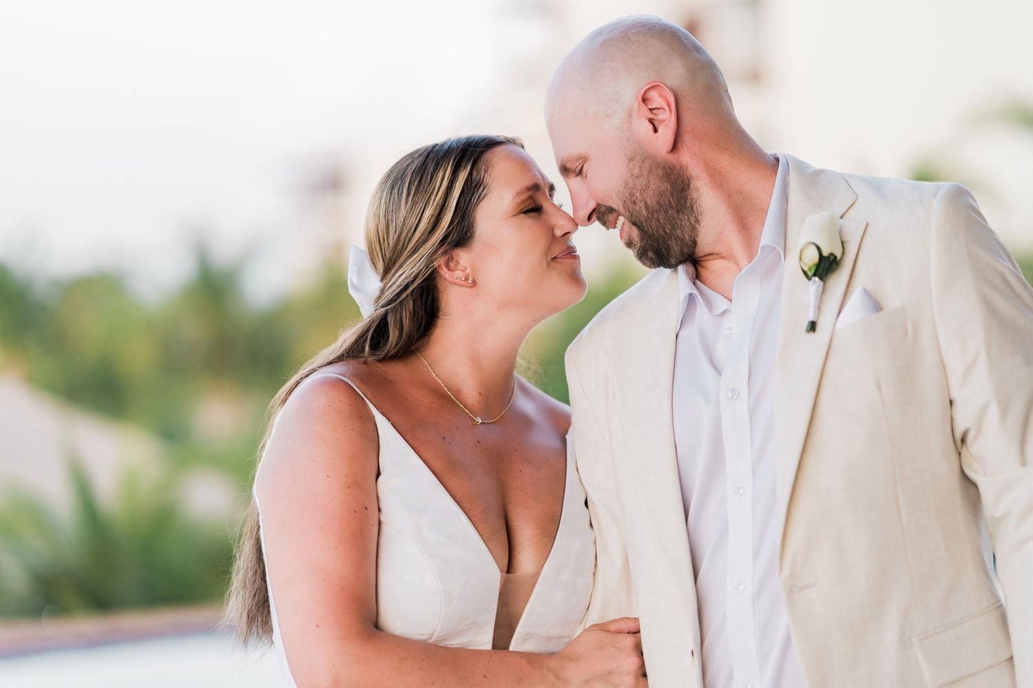 A bride and groom share a romantic nose-to-nose moment outdoors, dressed in a white gown and beige suit.