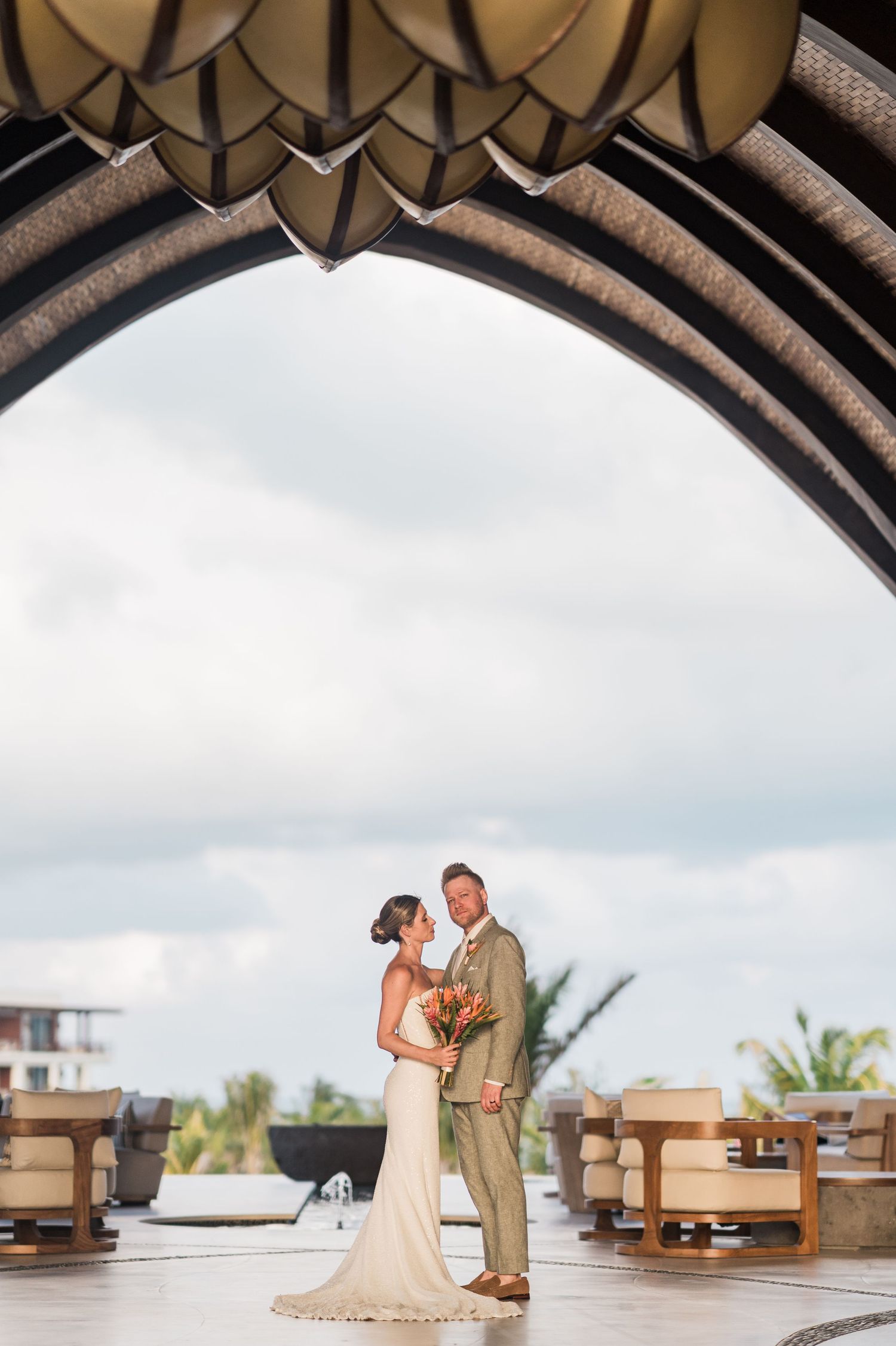 Bride and groom pose under elegant arch at luxury resort wedding venue with tropical backdrop.