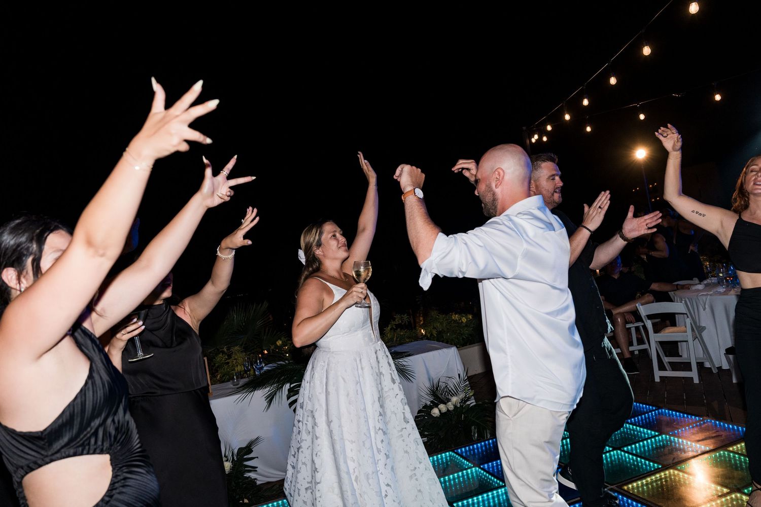 Bride and groom dancing joyfully with guests at outdoor wedding reception under string lights on colorful dance floor.
