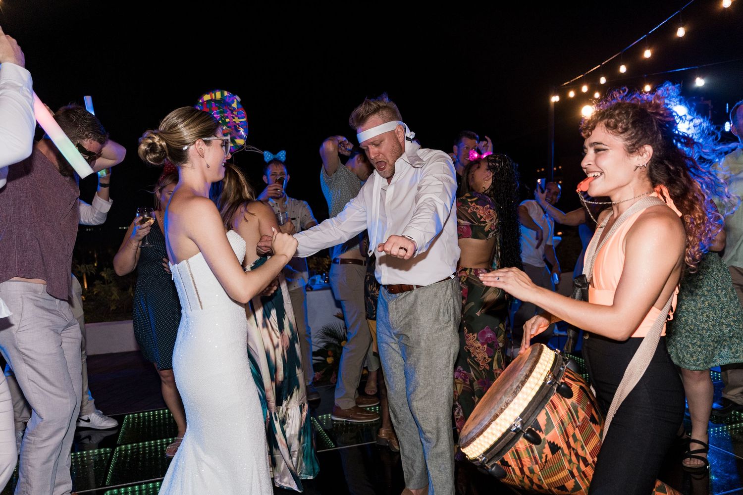 Bride and groom dancing joyfully at outdoor wedding reception while guest plays djembe drum at night.