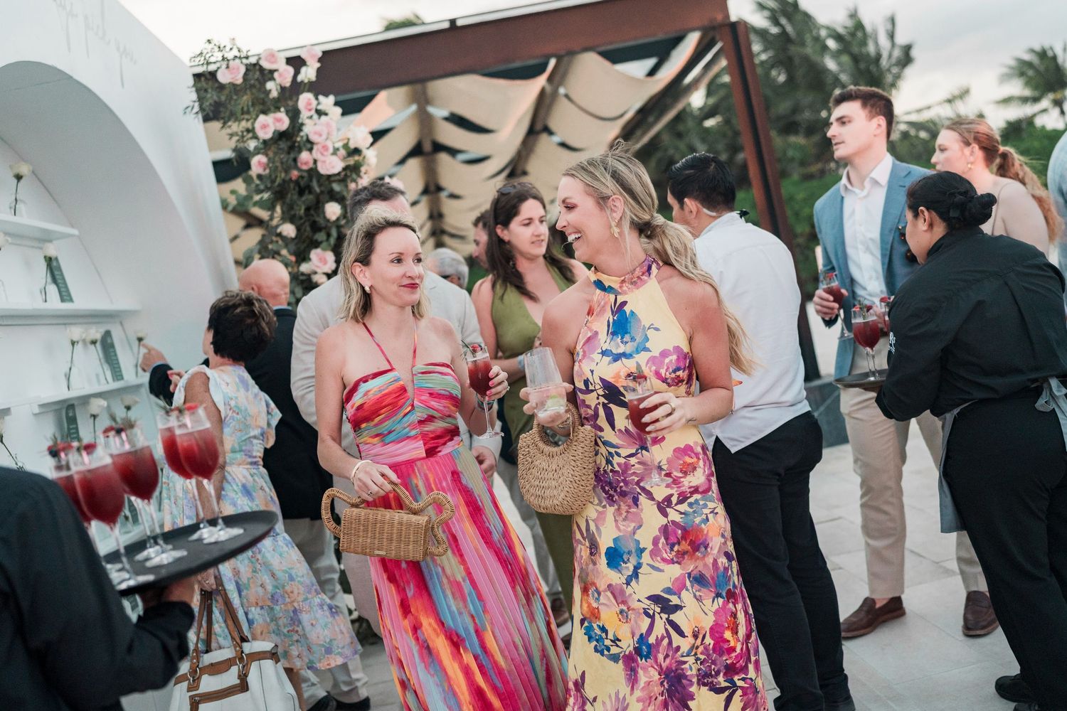 Guests in colorful floral dresses socializing at an elegant outdoor party, holding drinks and smiling.