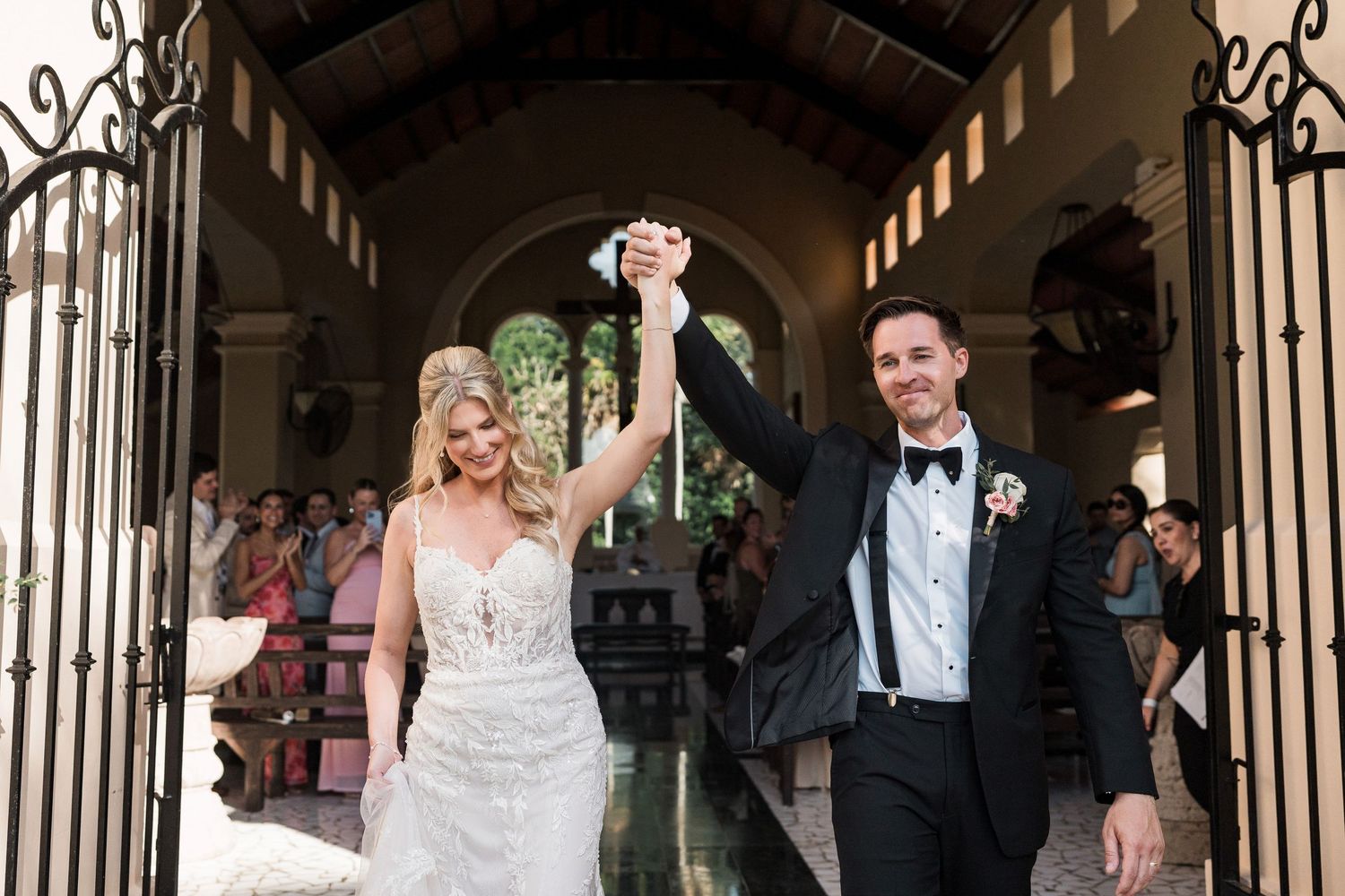 Joyful bride and groom raising hands at wedding ceremony exit, bride in lace gown, groom in black tuxedo.