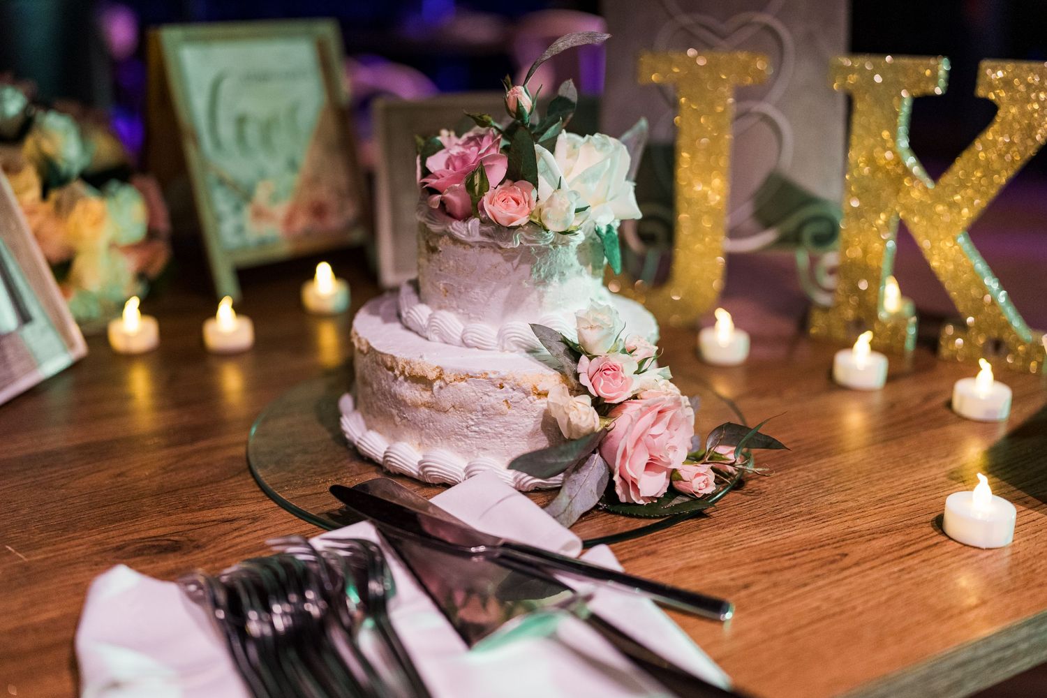 Elegant two-tier wedding cake decorated with pink and white roses surrounded by candles and gold initials.