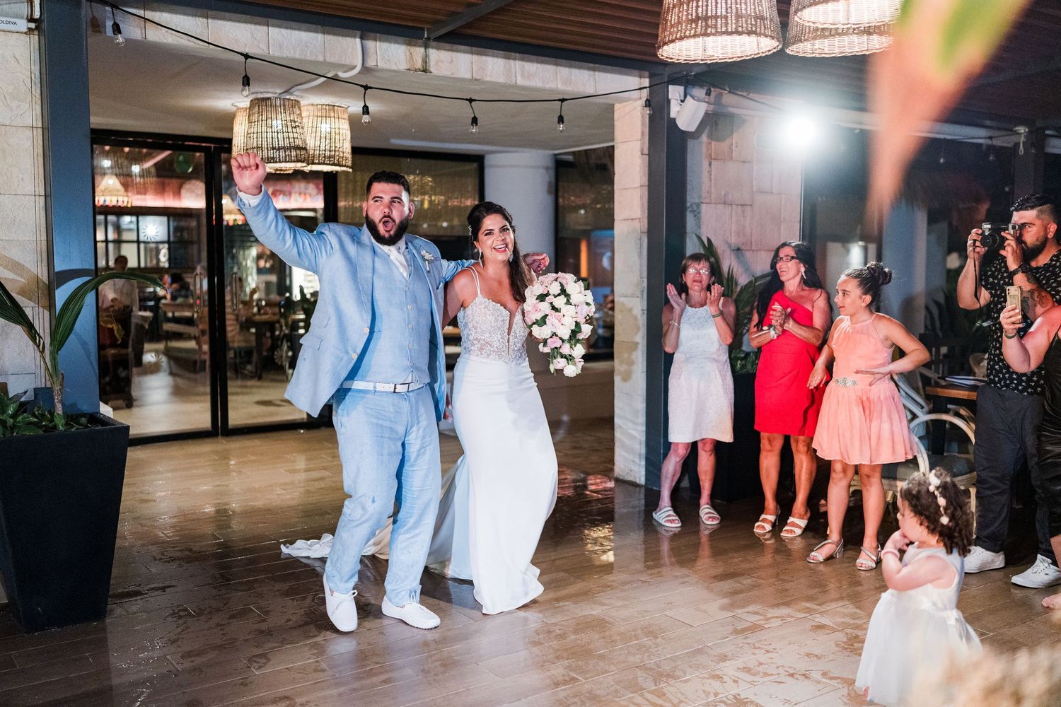 Bride and groom making grand entrance into wedding reception as guests cheer and applaud in festive venue.