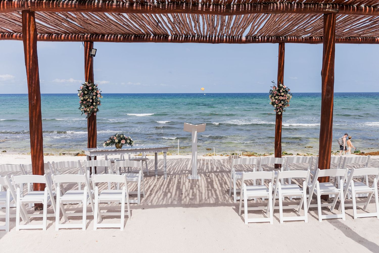 White chairs arranged for beach wedding ceremony under wooden pergola overlooking turquoise ocean.
