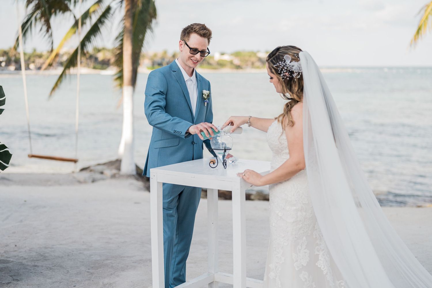 Groom and bride perform sand unity ceremony at white table on sunny beach wedding day.