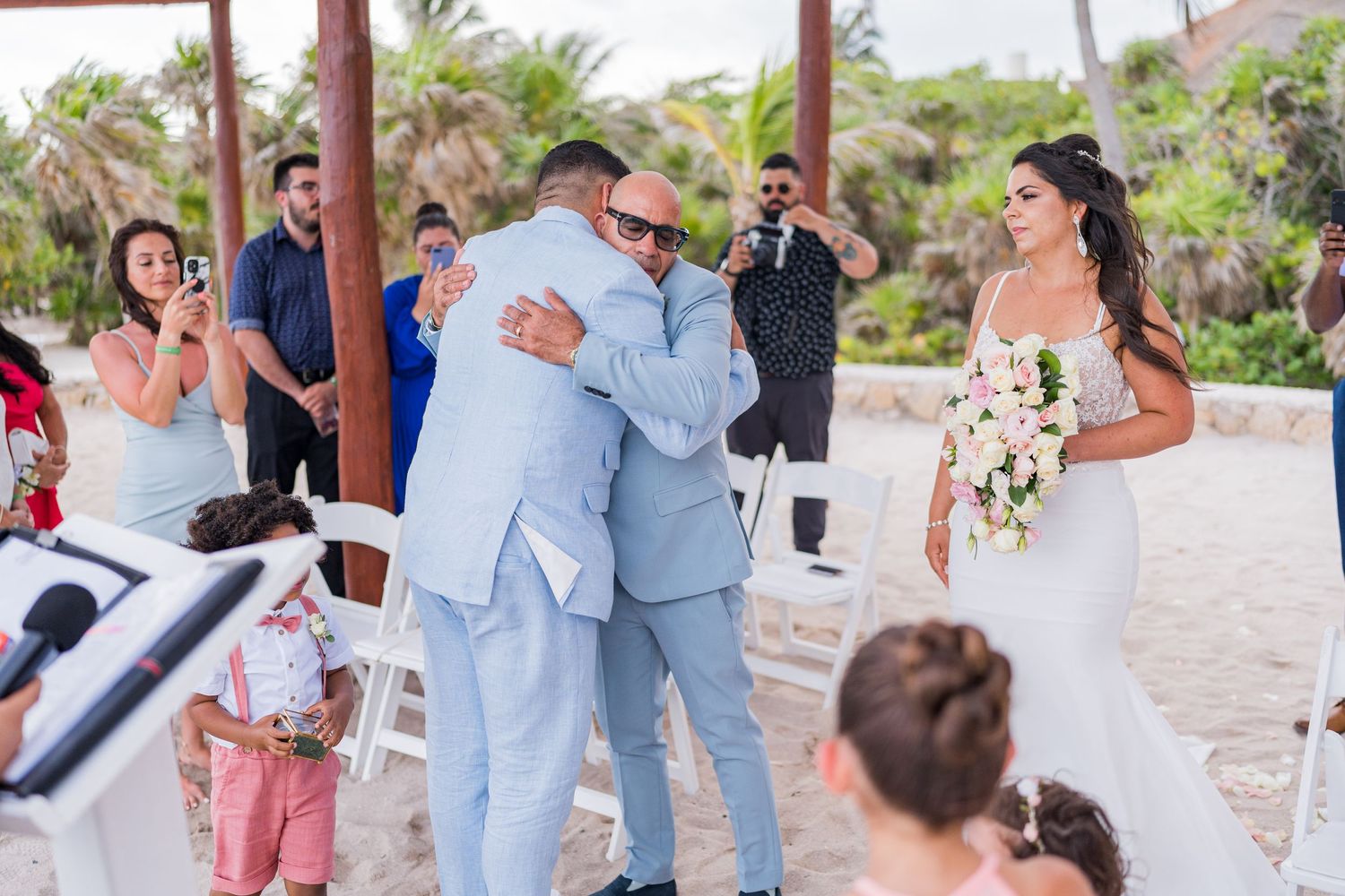 Groom hugging a guest at a beach wedding ceremony while the bride watches holding a bouquet.